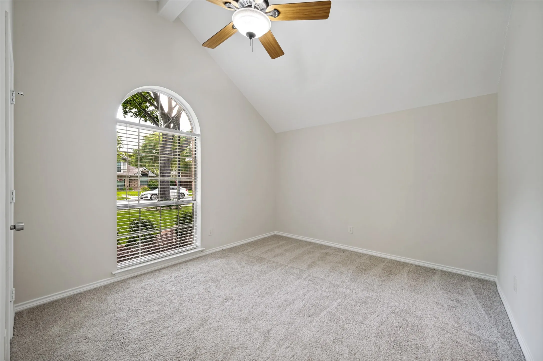 Arched window with street-facing views, vaulted ceiling, neutral wall paint, ceiling fan with light fixture, and gray carpet flooring