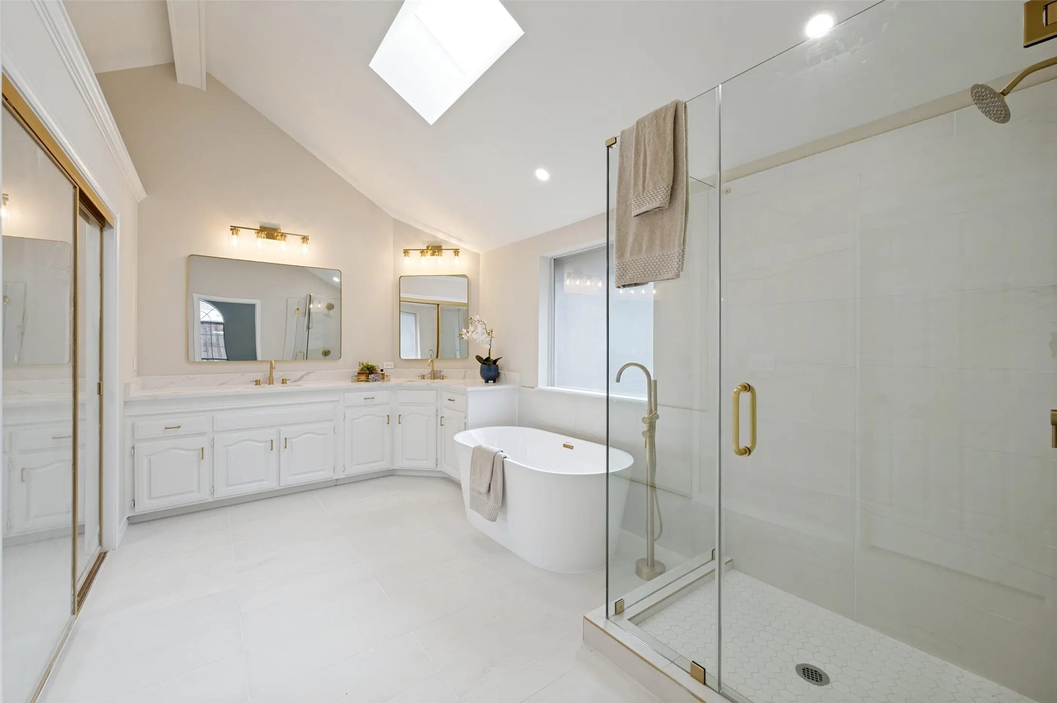 Vaulted ceiling bathroom featuring a large skylight, dual vanity with white cabinetry, freestanding soaking tub, and a glass-enclosed shower with gold-tone fixtures