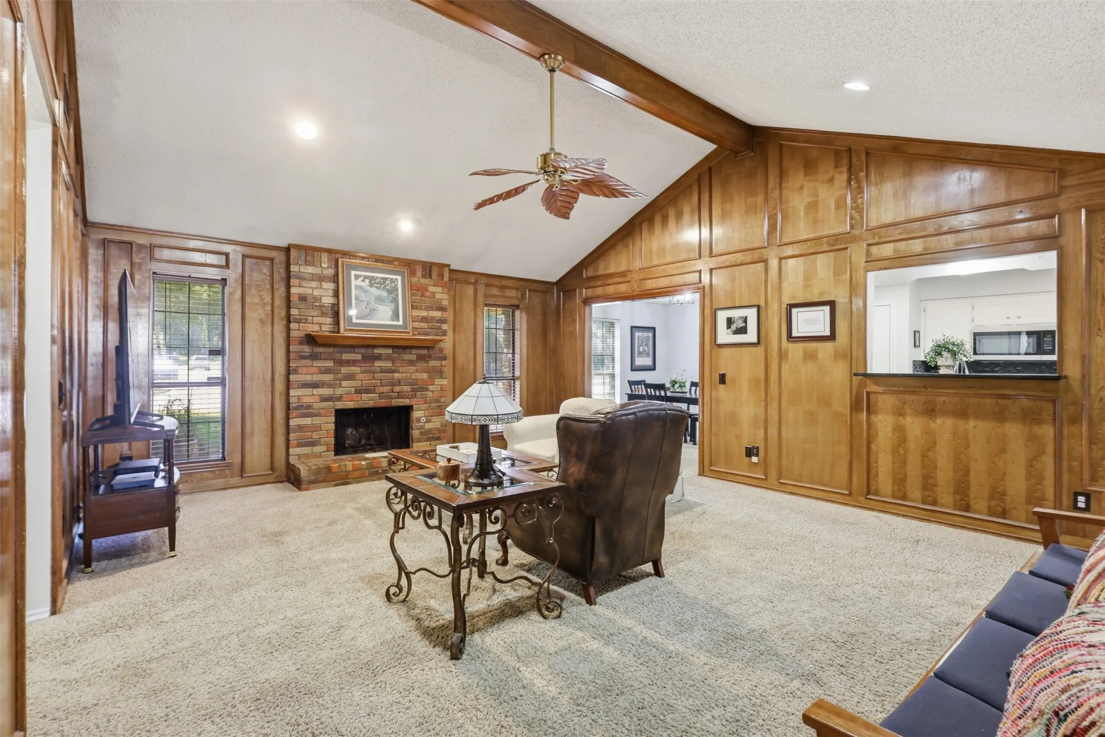 Spacious living area featuring a vaulted ceiling with exposed wood beams, a brick-front fireplace, wood paneling, and an interior pass-through window