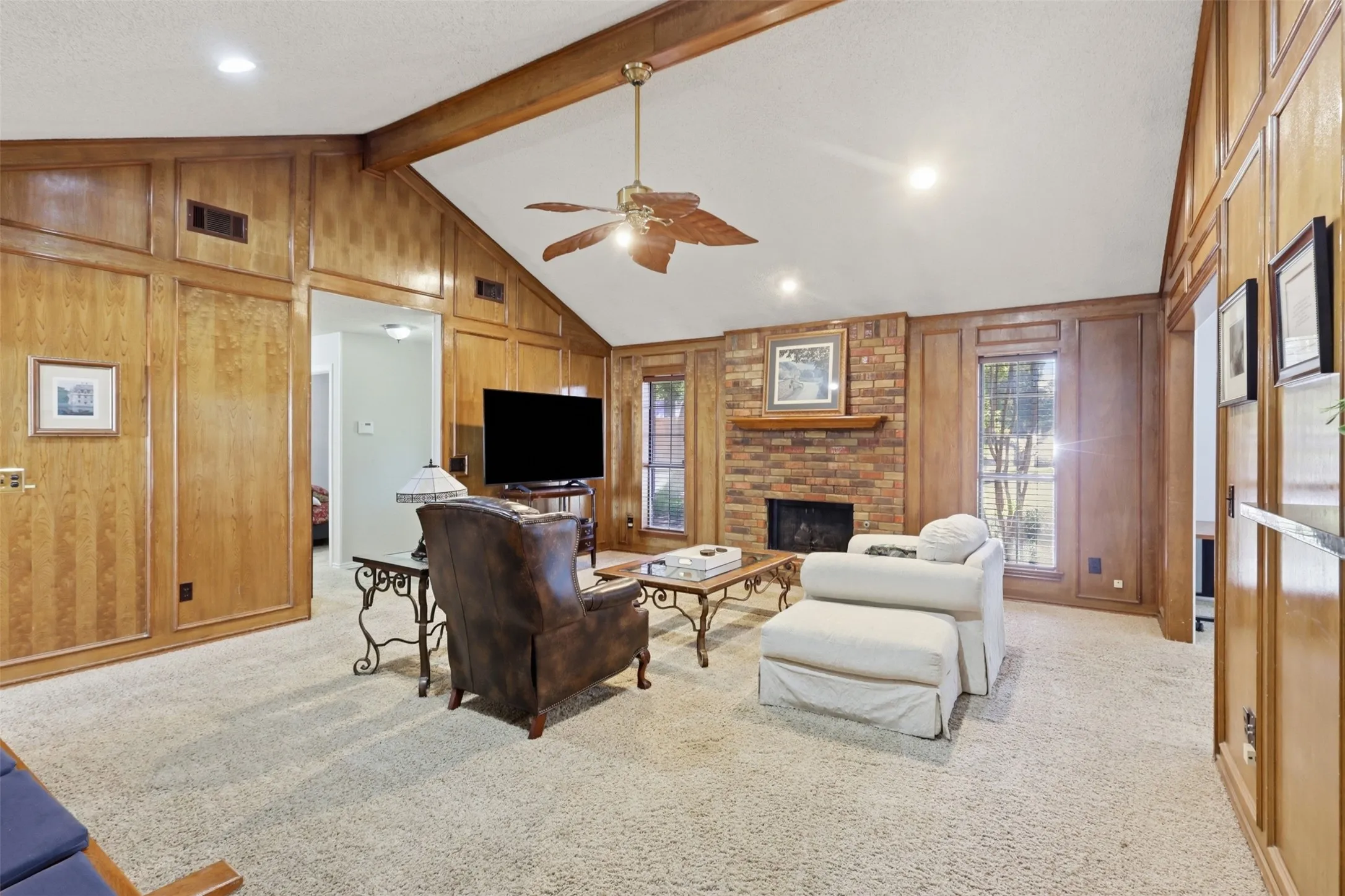 Vaulted ceiling with exposed wood beams, brick fireplace with wood mantel, wood paneling, recessed lighting, and a ceiling fan