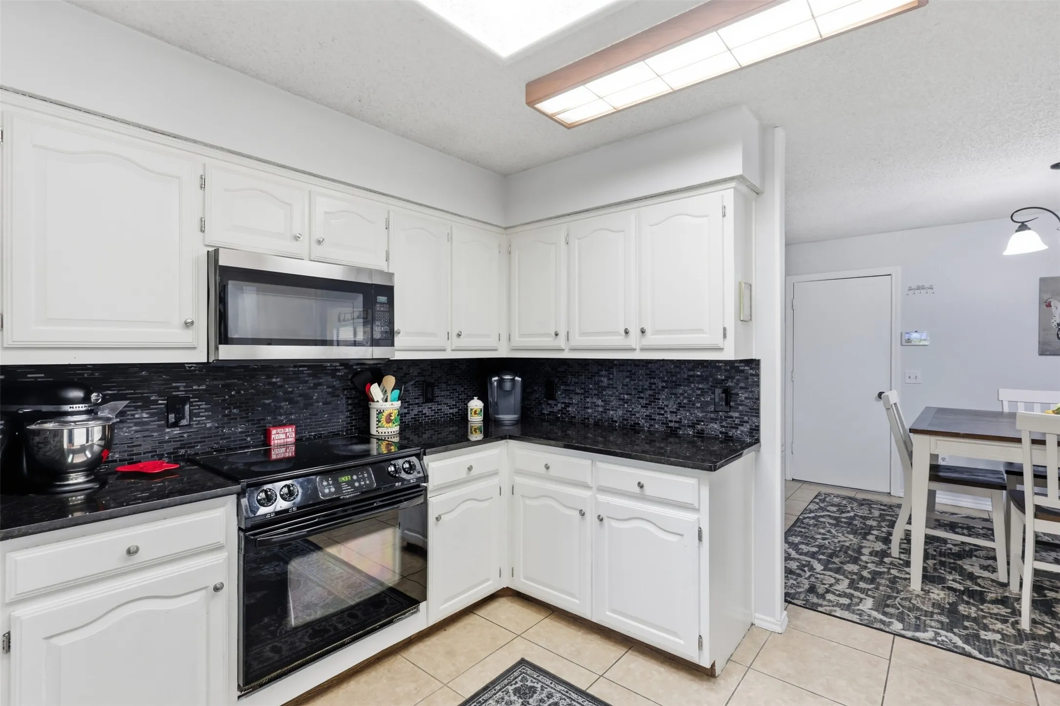 Kitchen featuring white cabinetry, black stone countertops, a coordinating black tile backsplash, and stainless steel microwave