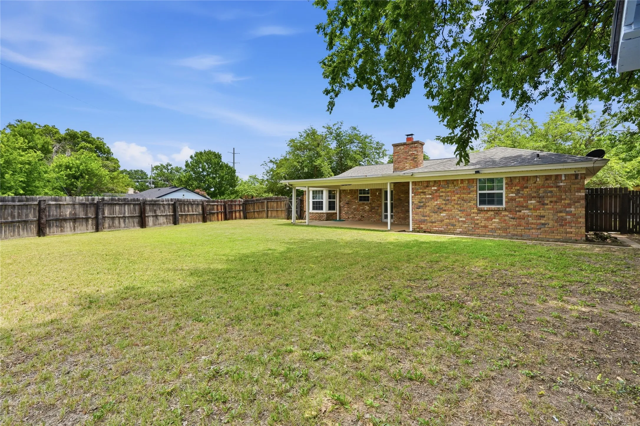 Expansive grassy backyard featuring a full privacy fence, brick exterior with multiple windows, covered patio, and a brick chimney
