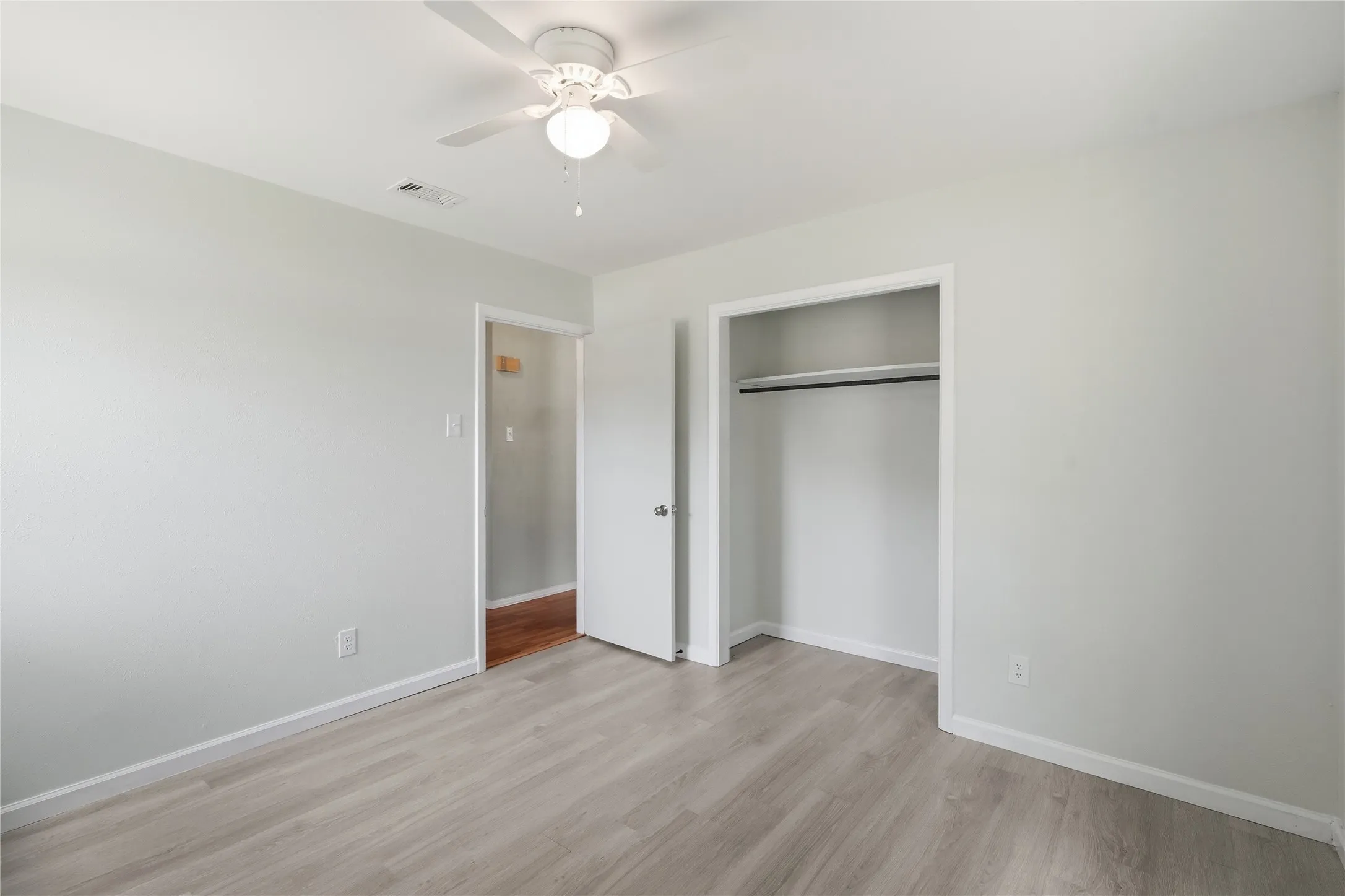 Bright room featuring light-toned walls, wood-finish flooring, a ceiling fan with integrated lighting, and an open closet with a hanging rod
