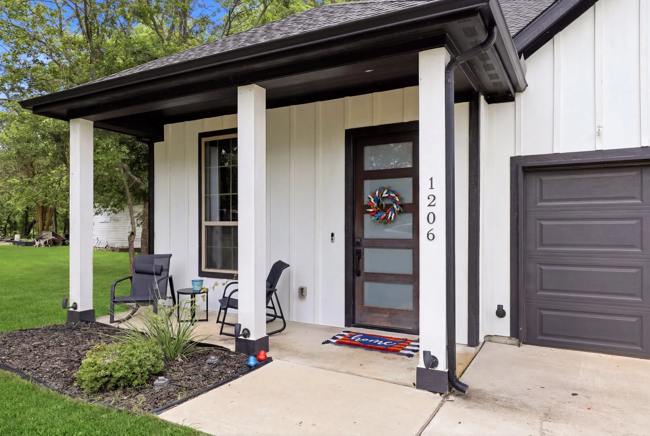 Contemporary exterior featuring white vertical siding, a dark wood-finish entry door with frosted glass panels, a covered front porch with white columns, and a paneled garage door
