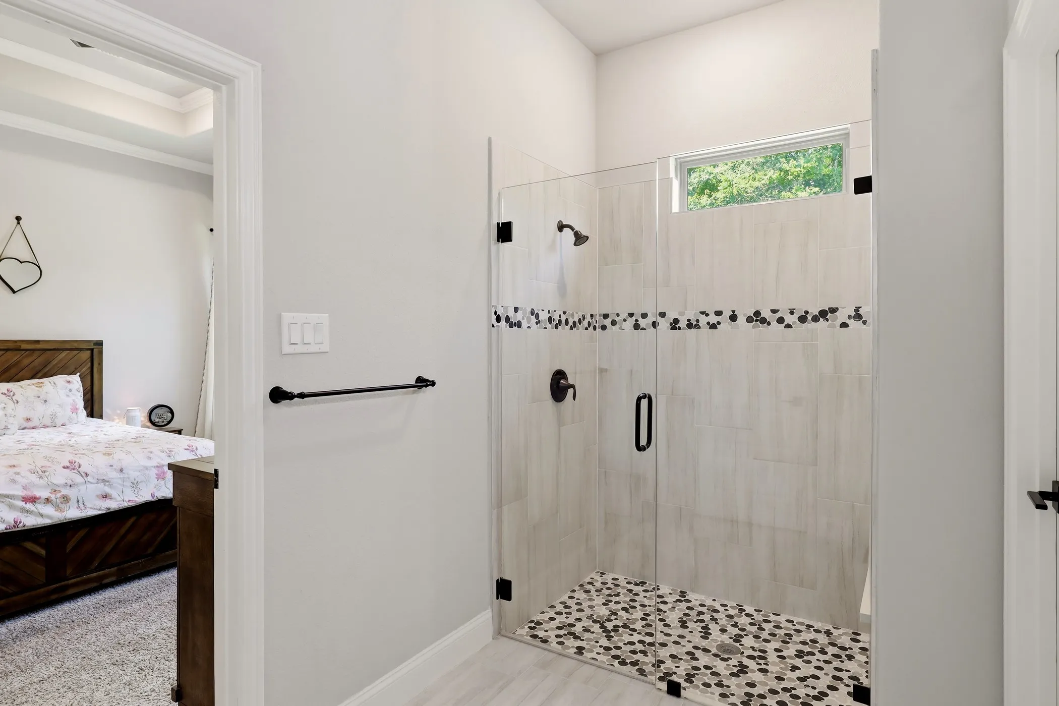 Glass-enclosed shower featuring light-toned tile walls with a decorative pebble accent band, a pebble-tiled shower floor, and a transom window