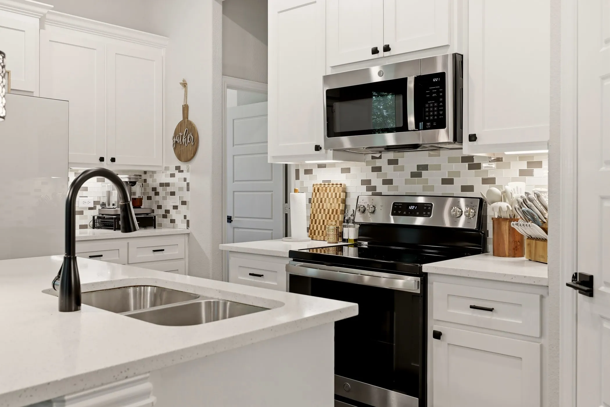 Modern kitchen featuring white shaker cabinetry, stainless steel appliances, a multi-tone tile backsplash, light-toned countertops, and a dual basin undermount sink with a black gooseneck faucet