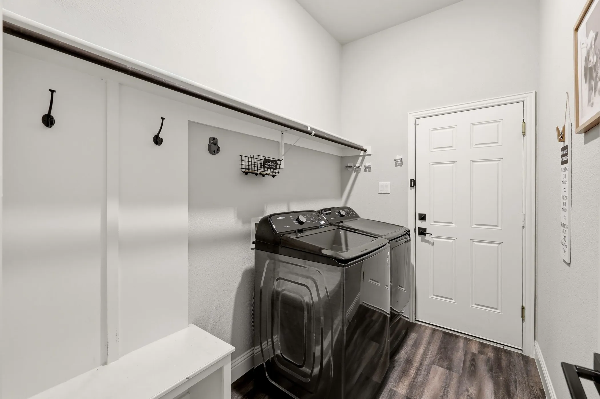 Laundry room featuring wood-finish flooring, built-in shelving, white paneled walls with hooks, and a white paneled door with matte black hardware