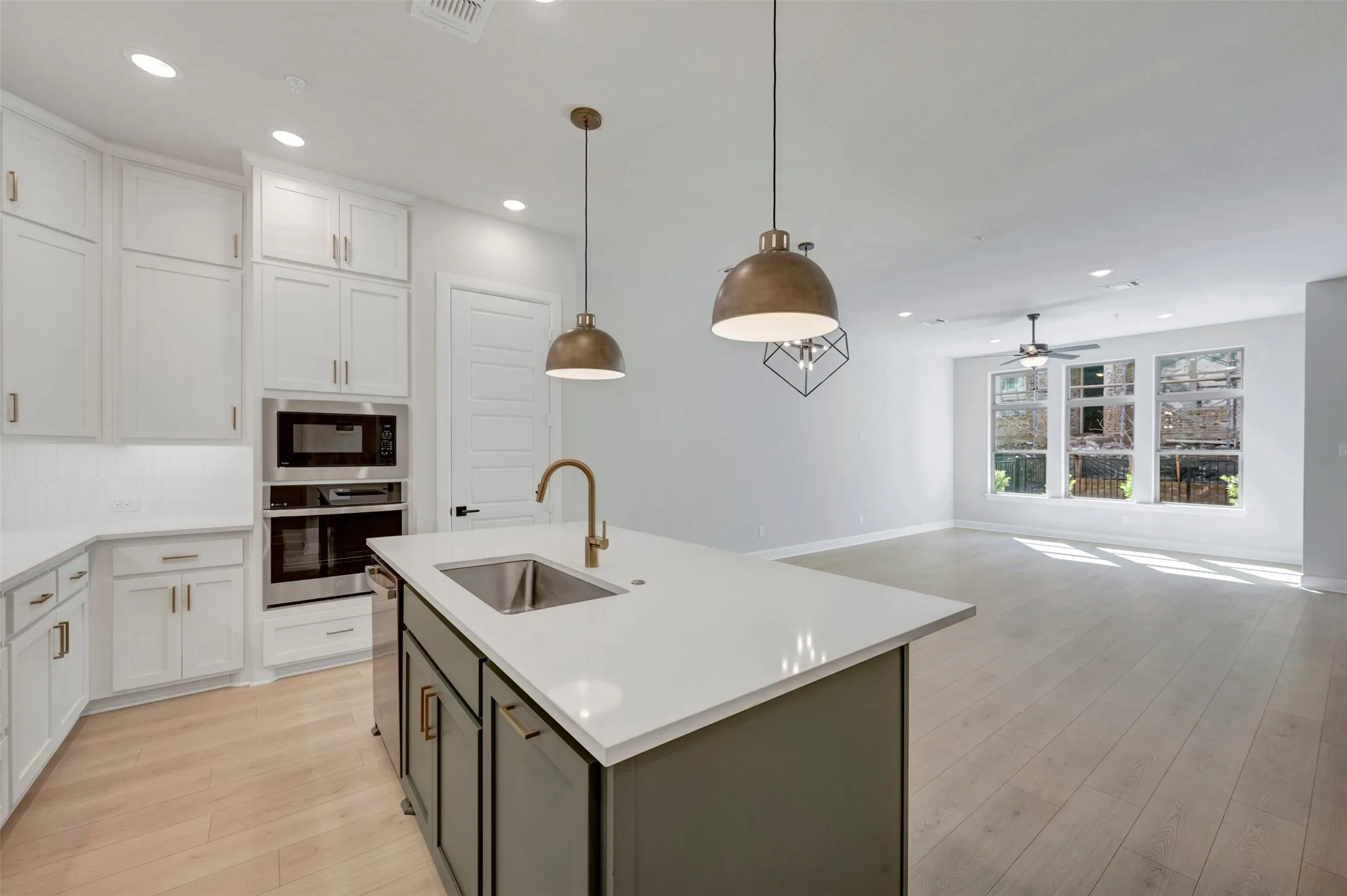 Kitchen island with an undermount sink and a brass-finish faucet, white shaker-style cabinetry, built-in stainless steel appliances, light wood-finish flooring, and multiple windows with white trim