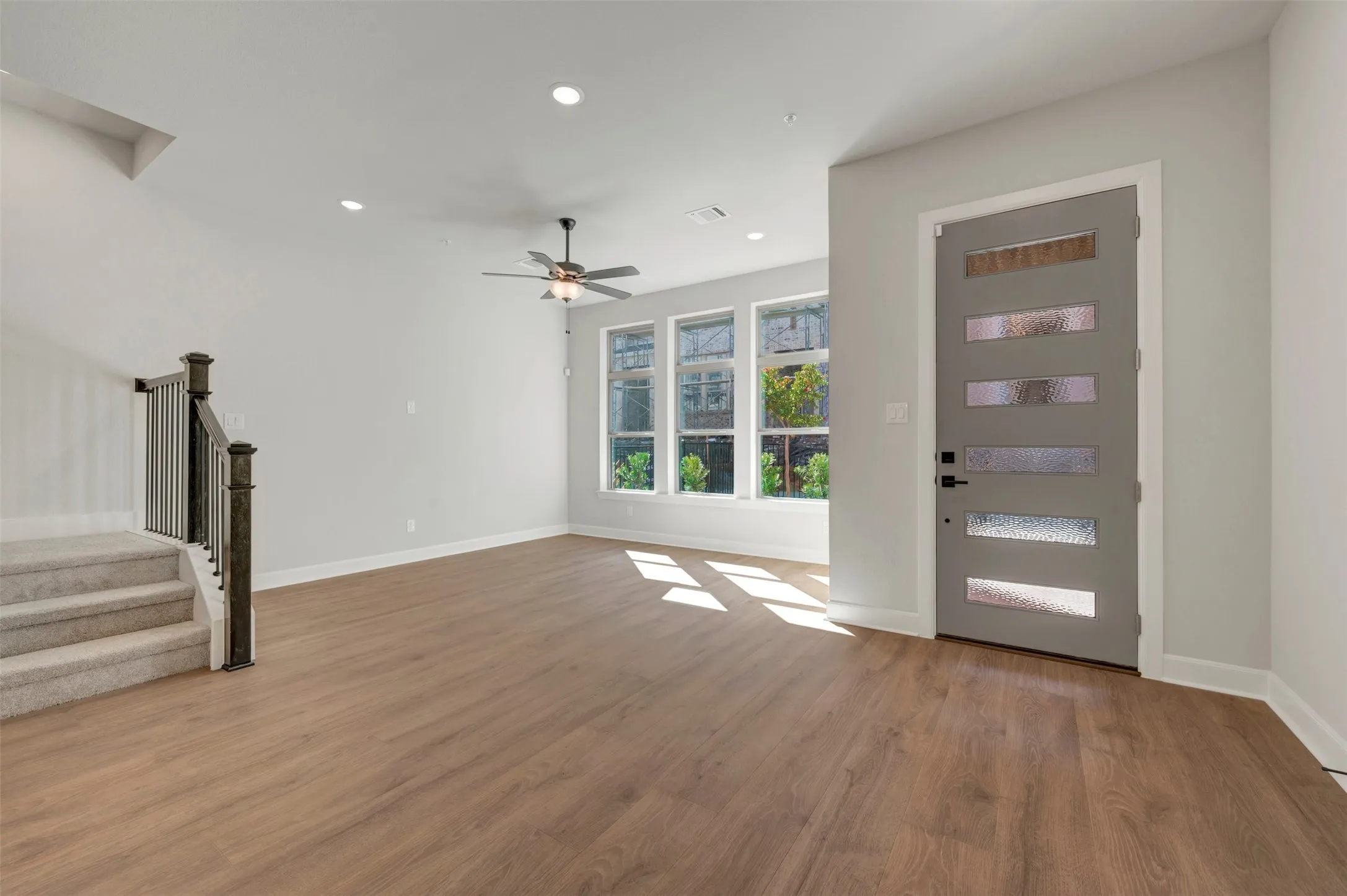 Wood-finish flooring throughout, modern entry door with frosted glass panels, recessed lighting, ceiling fan, and a carpeted staircase with dark wood-finish newel posts and metal balusters