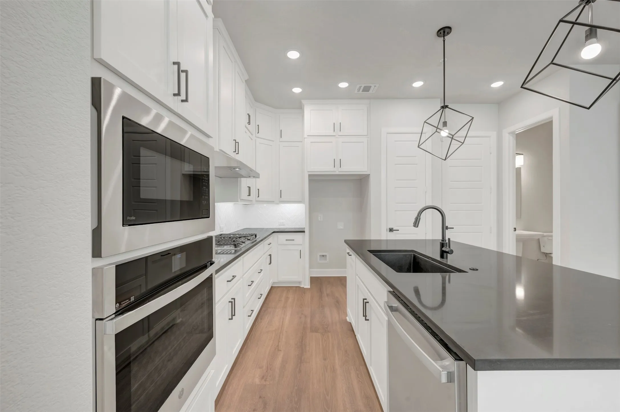 Kitchen featuring a central island with a built-in sink, white cabinetry with dark hardware, stainless steel appliances, wood-finish flooring, and recessed lighting