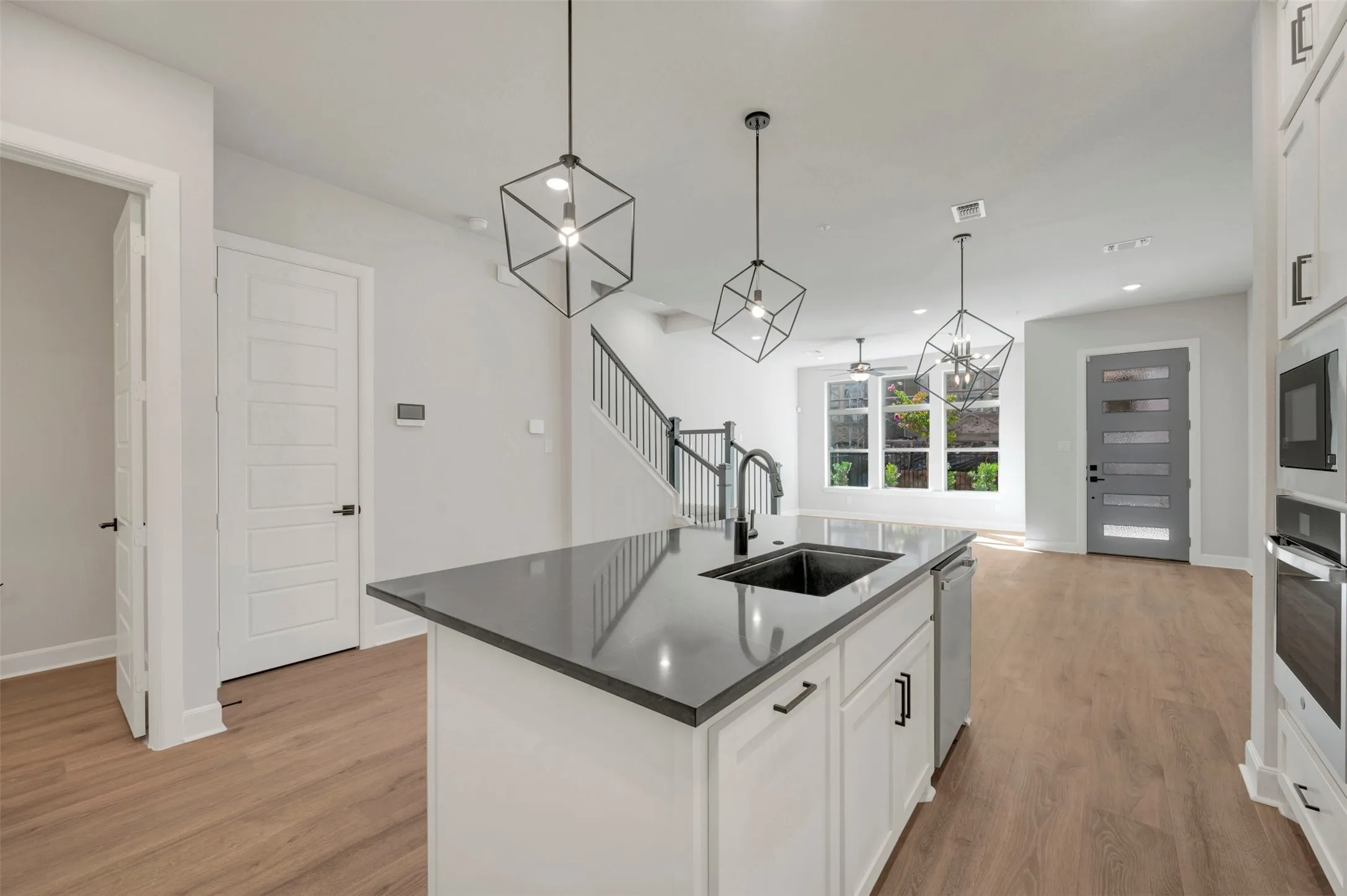 Modern kitchen featuring a large center island with black countertop and integrated sink, white cabinetry with hardware accents, stainless steel appliances, pendant lighting with geometric frames, and wood-finish flooring throughout