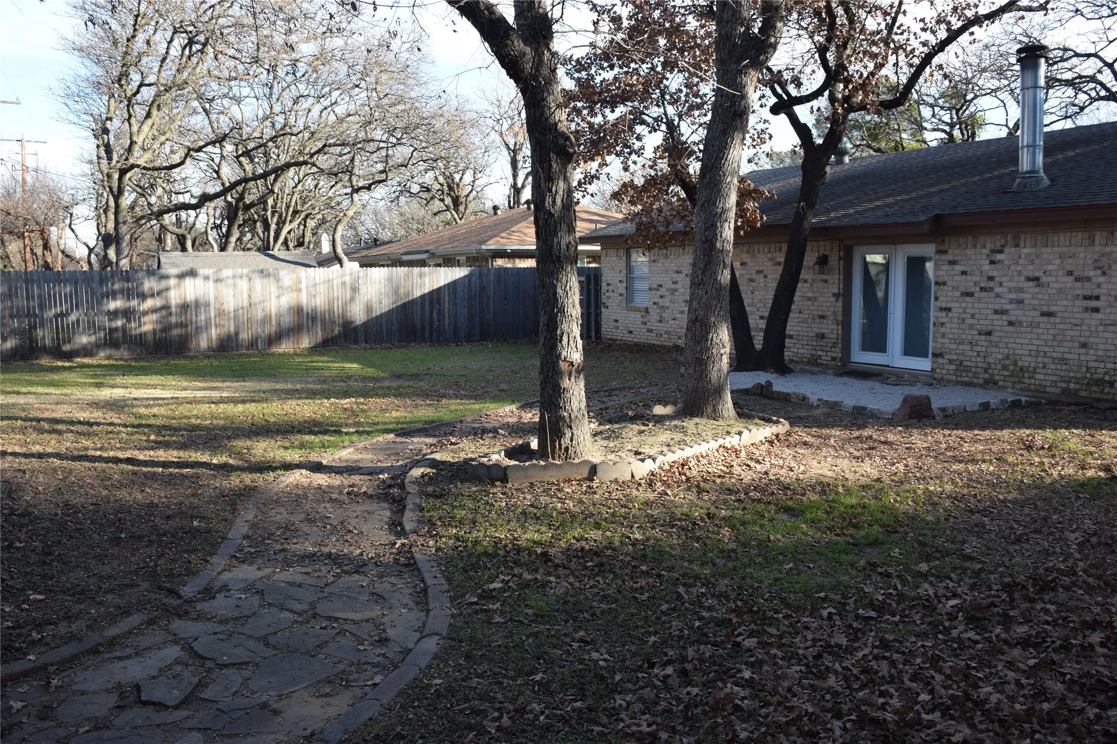 Expansive backyard featuring a stone pathway, mature trees, wood fence, and a brick exterior with white double doors opening onto a patio