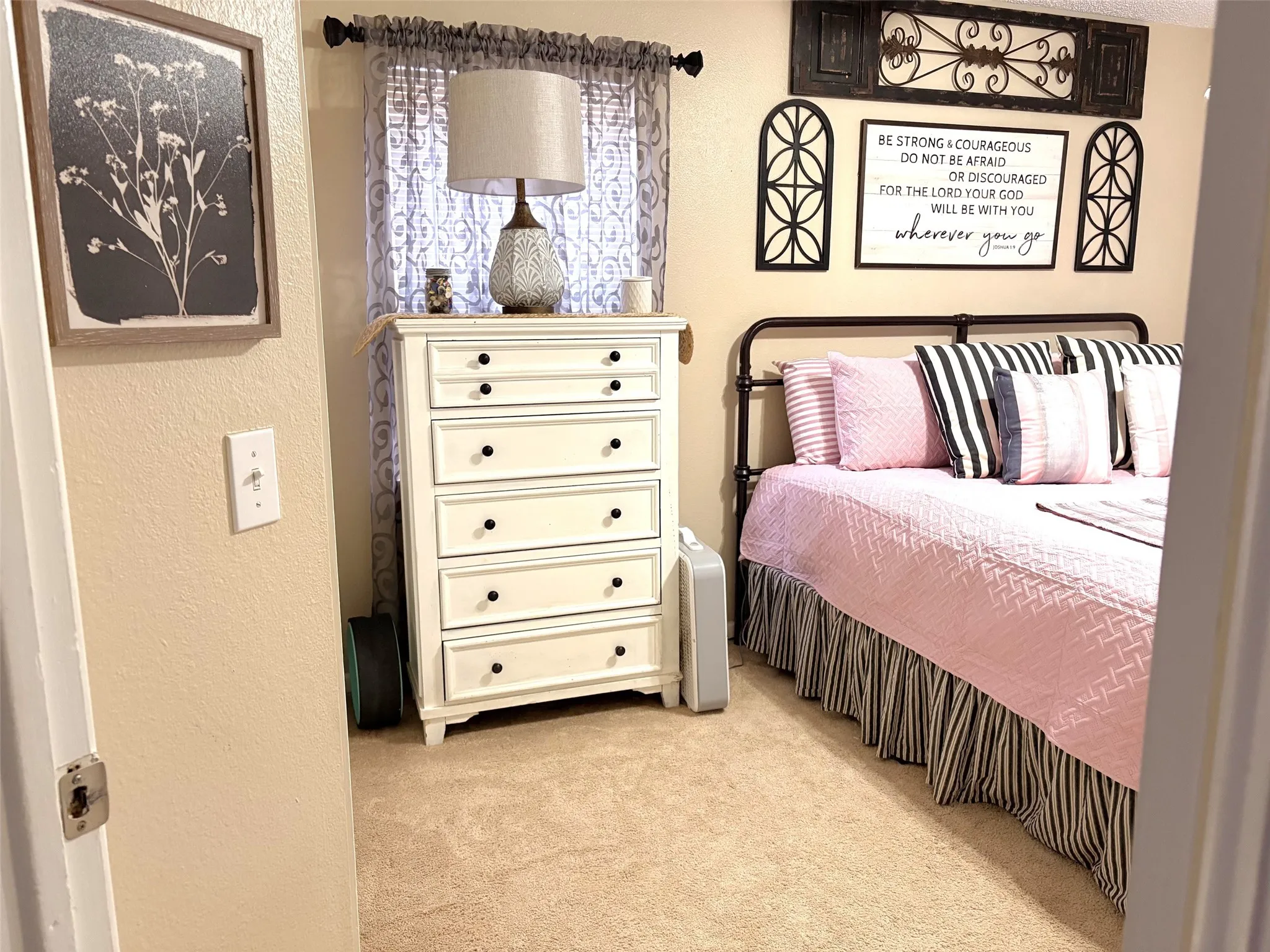Carpeted room featuring a window with patterned drapes, a decorative metal headboard, and neutral-toned walls