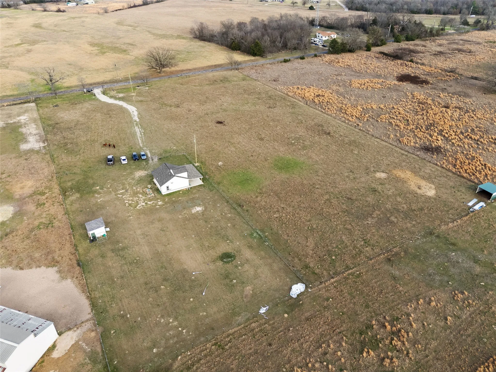 Expansive acreage featuring a single-story residence with a gray shingled roof, a smaller auxiliary building, a separate green-roofed structure, and a long gravel driveway