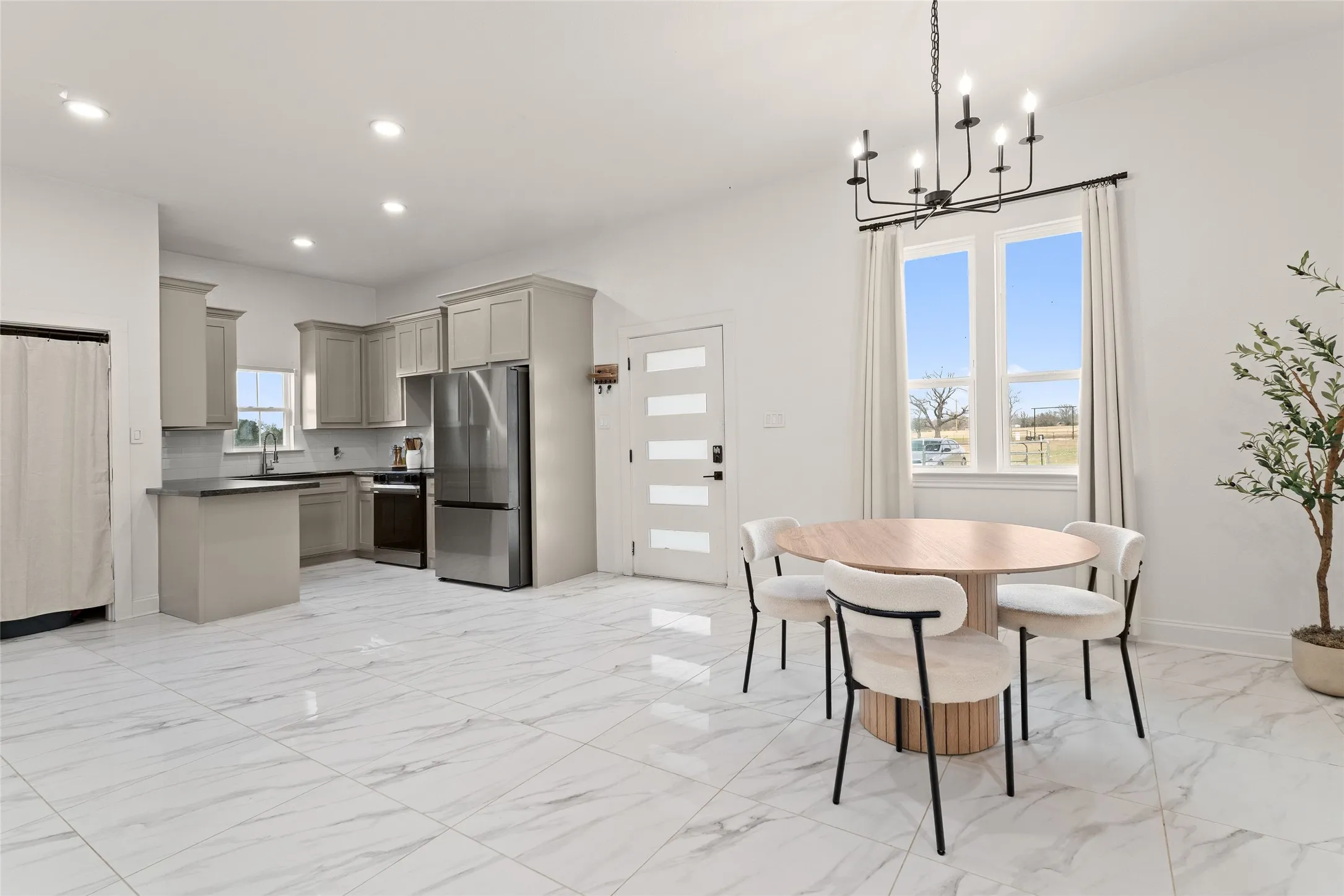 Open-concept living space featuring large format marble-look tile flooring, a modern kitchen with grey cabinetry and stainless steel appliances, and a dining area with a contemporary black chandelier