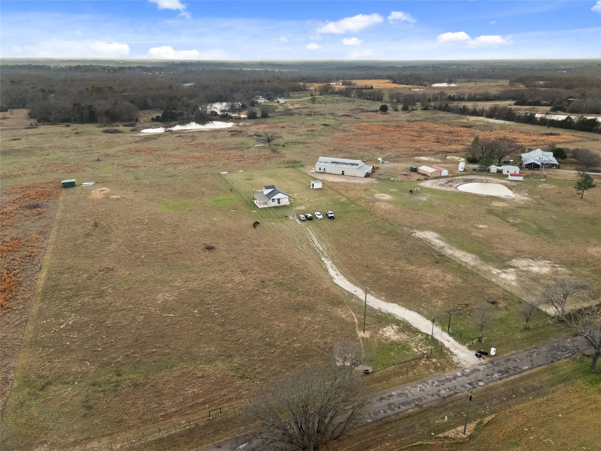 Expansive rural property featuring multiple structures, including a primary residence with a dark roof and white siding, a large detached outbuilding with a light-colored roof, and additional smaller outbuildings