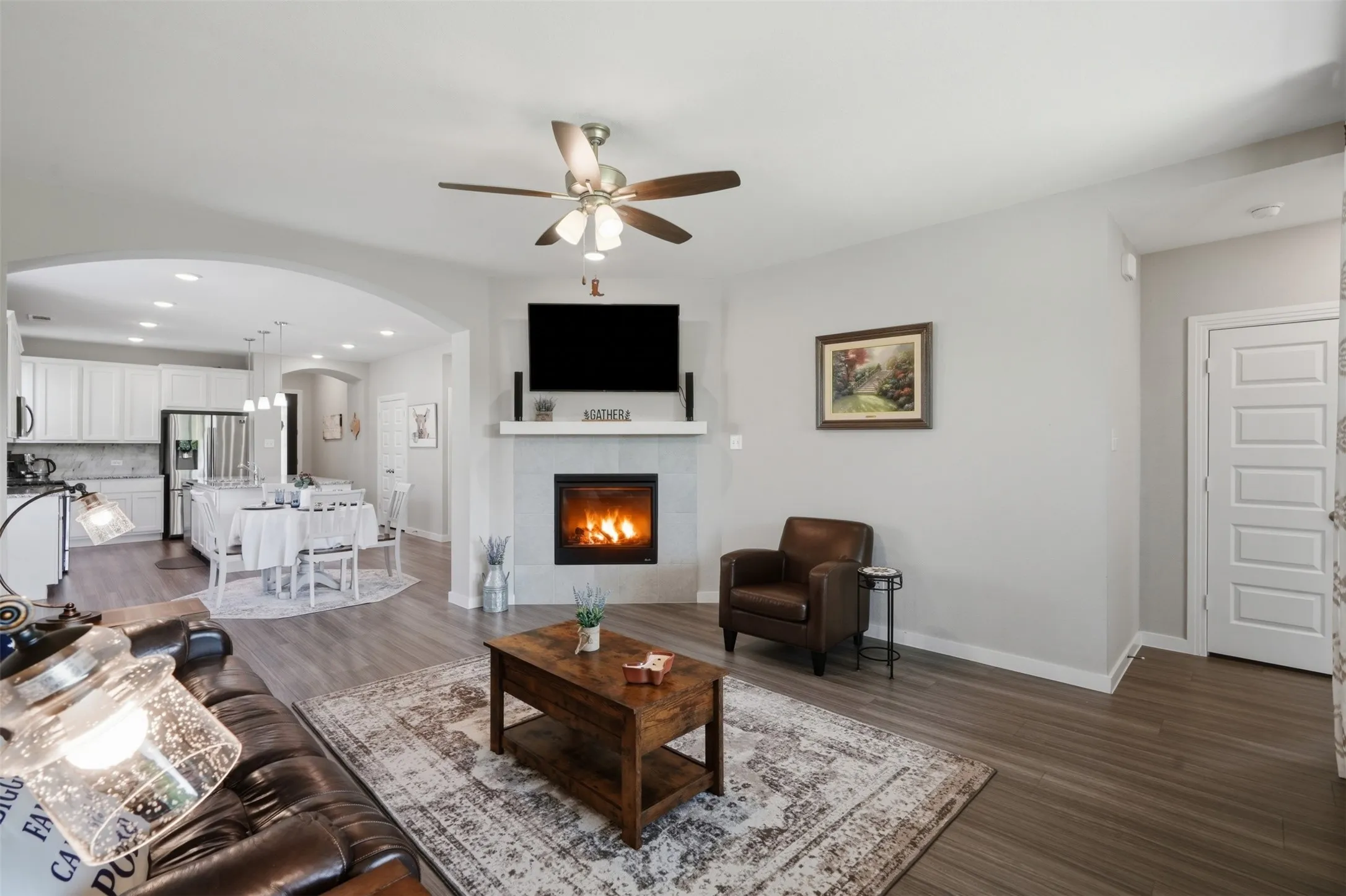 Well connected living room with a tile fireplace, wall mounted TV, ceiling fan, and an elegant arched opening leading to the kitchen and dining room.