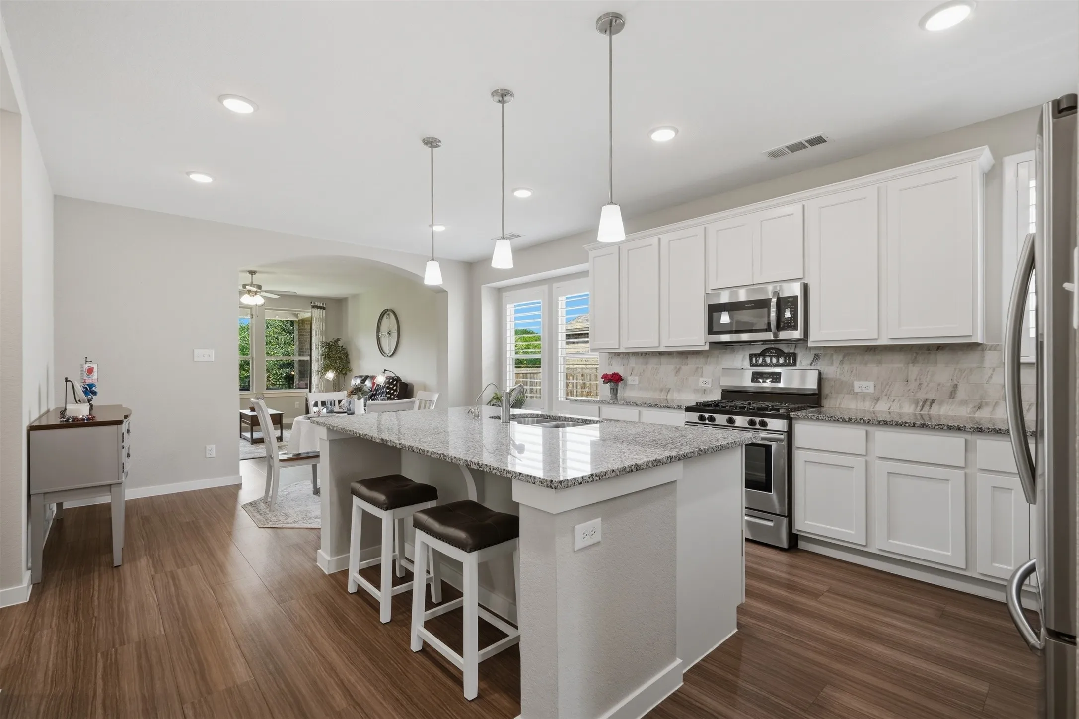 Spacious kitchen with white cabinets, granite island, stainless steel appliances, tile backsplash, pendant lights, and an open view to the living area.