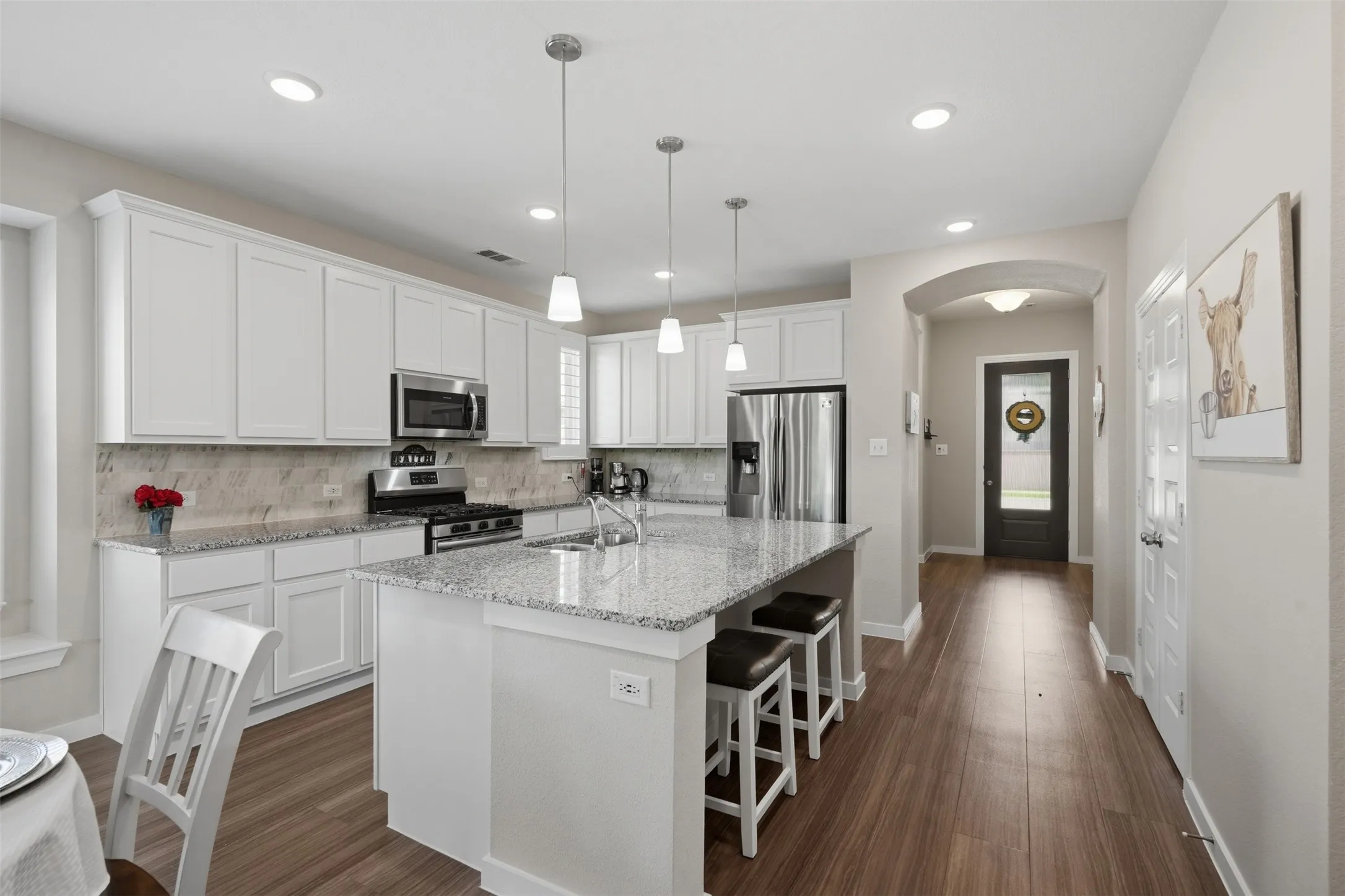 Open kitchen with granite island, bar seating, white cabinets, gas range, stainless steel appliances, tile backsplash, pendant lights, and an arched hallway entry.
