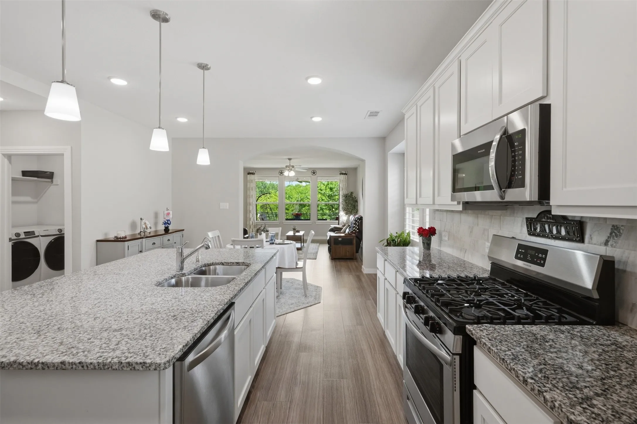 Functional kitchen featuring granite island with double sink, gas range, stainless steel appliances, tile backsplash, pendant lights, and direct access to the laundry room.