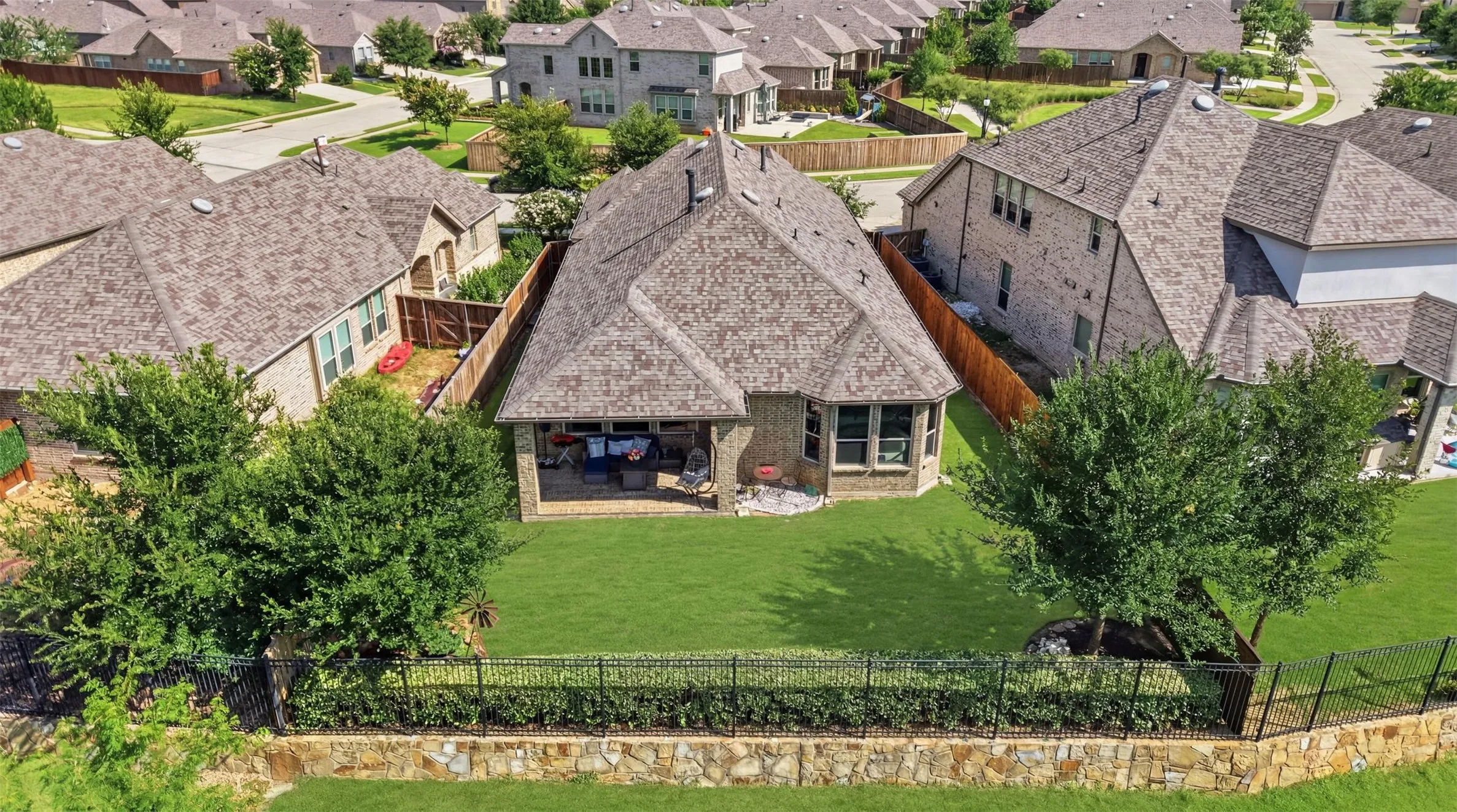 Bird's-eye view showcasing a generously sized backyard with a covered patio, manicured lawn, mature shade trees, and a stone perimeter fence.