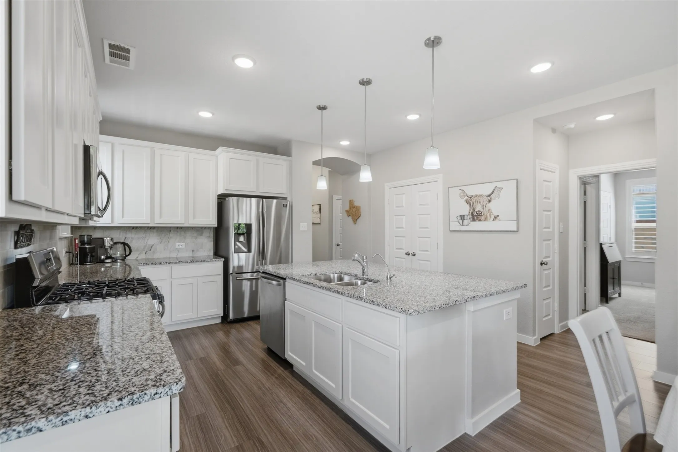 Bright open kitchen with granite island, white cabinetry, tile backsplash, stainless steel appliances, pendant lighting, and wood look plank flooring.
