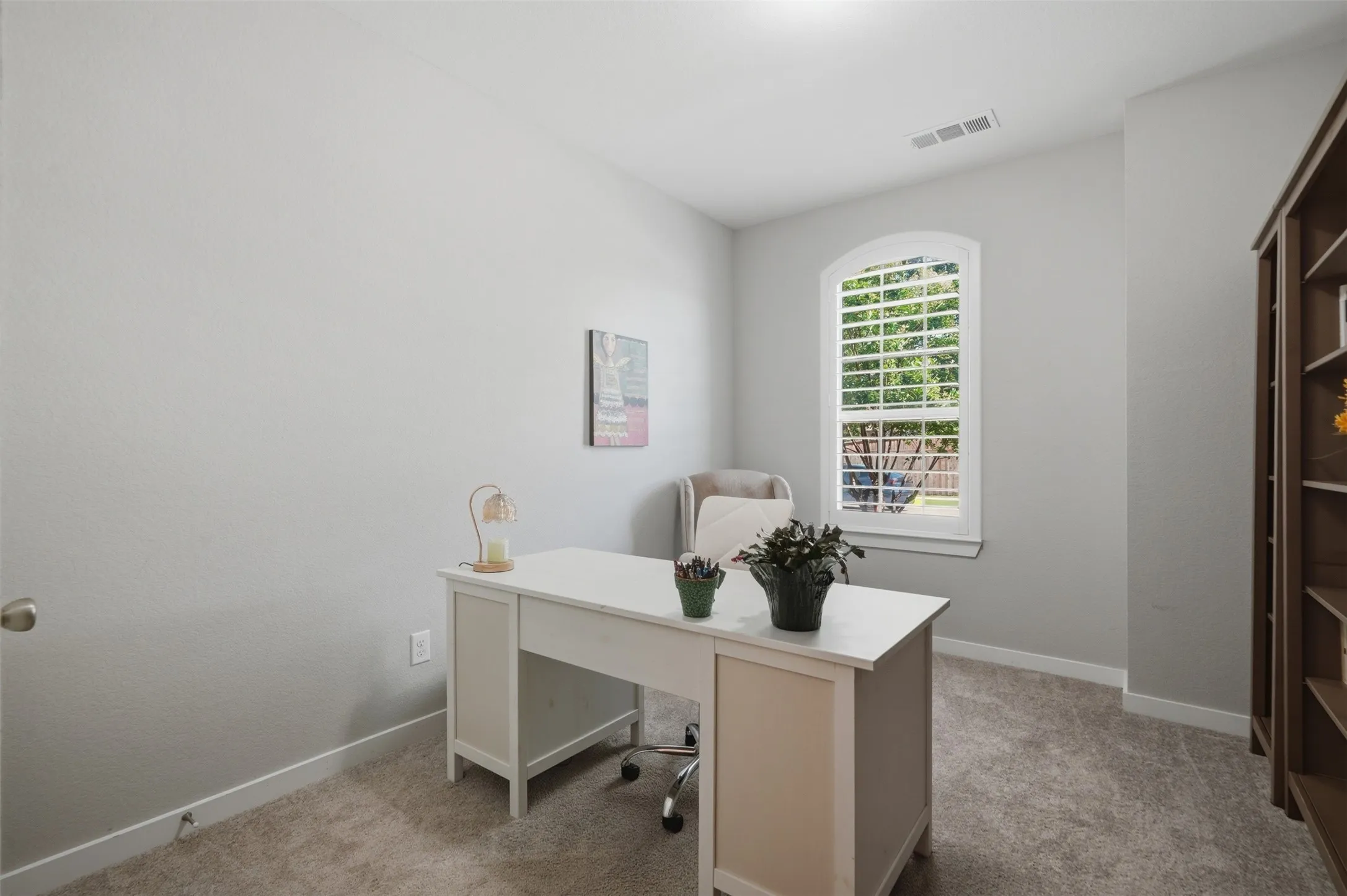 Home office space featuring plush carpet, neutral walls, plantation shutters, and a bright arched window overlooking the yard.