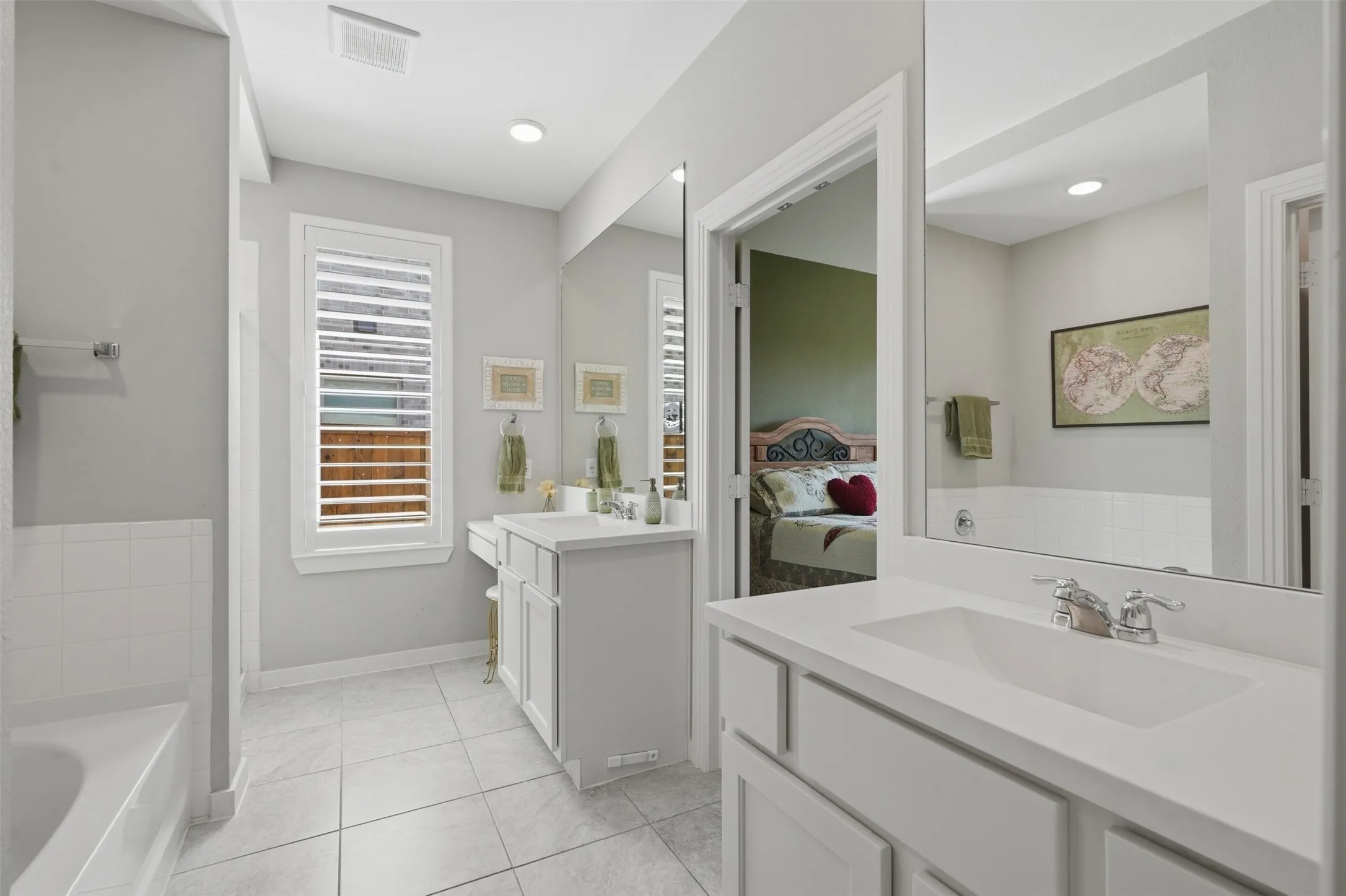 Primary bathroom boasting dual vanities with white cabinetry, large mirrors, soaking tub, tile flooring, and plantation shutters.