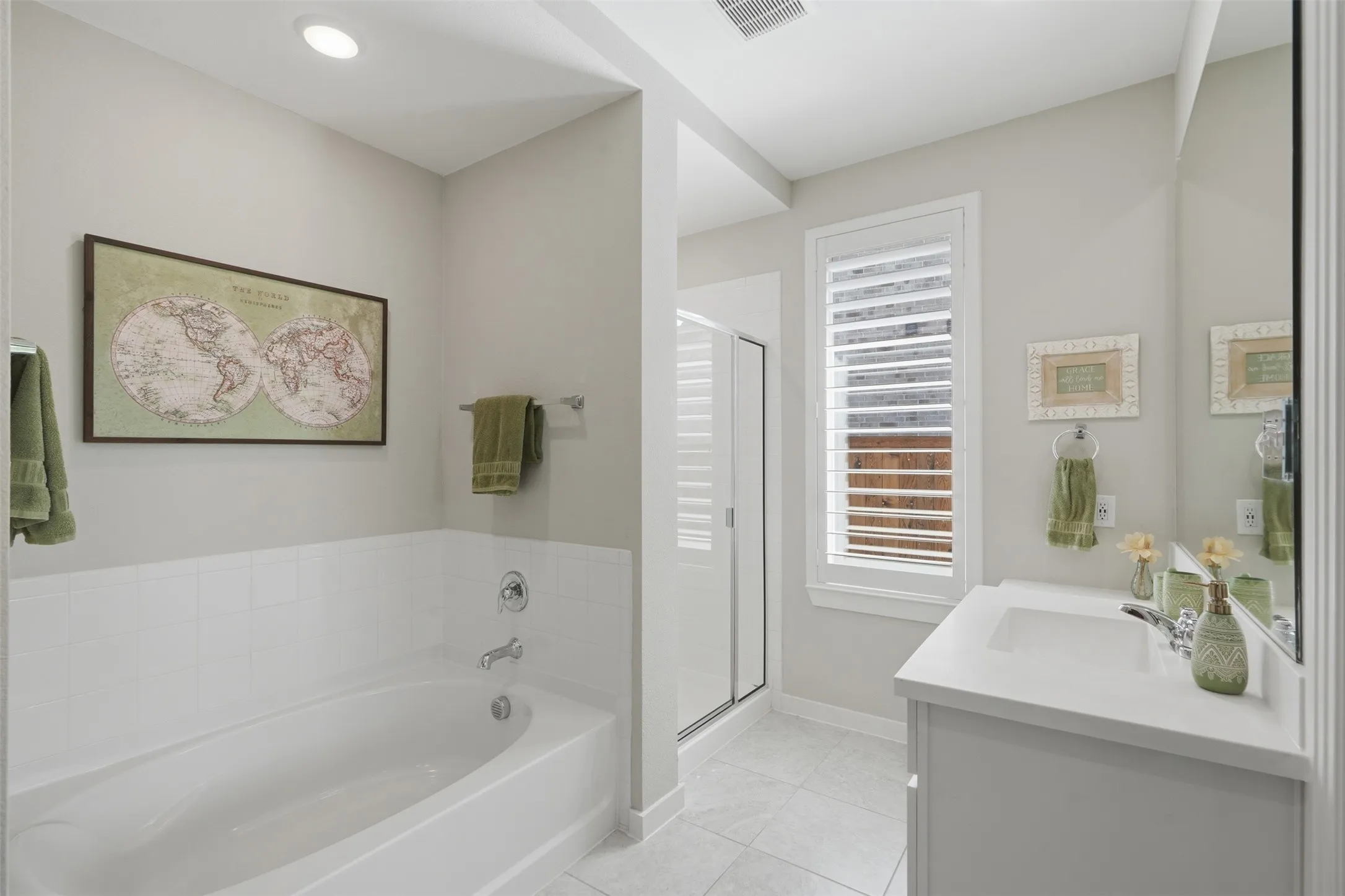Clean and bright primary bathroom featuring a soaking tub, separate walk-in shower, white vanity, tile flooring, and plantation shutter window