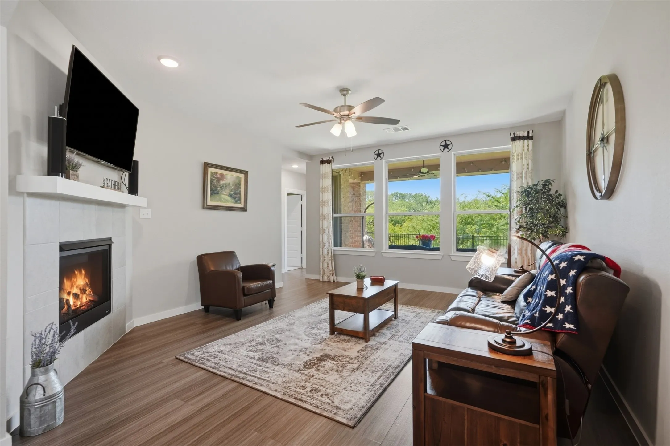 Spacious living room featuring a tile surround fireplace, wood look floors, ceiling fan, and large windows with greenbelt views.