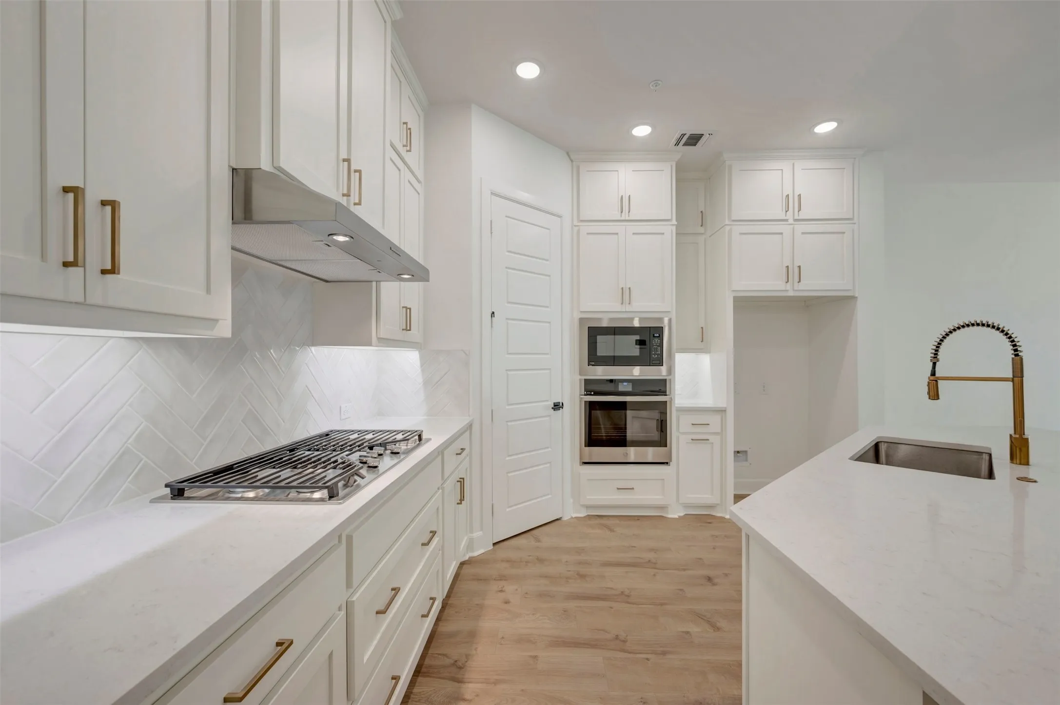 White shaker cabinetry with gold-finish hardware, stainless steel gas range, white herringbone tile backsplash, built-in microwave and oven, and recessed lighting