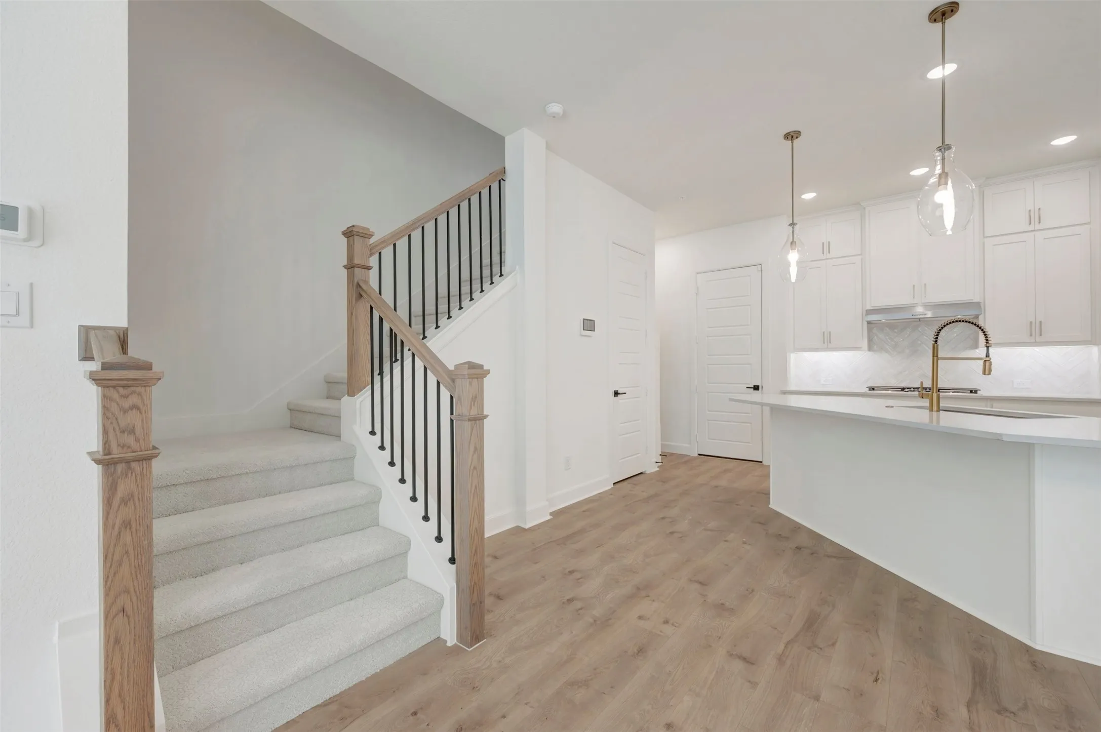Wood-finish flooring, staircase with natural wood handrails and black iron spindles, white kitchen cabinetry, kitchen island with integrated sink and gold-finish faucet, and pendant lighting