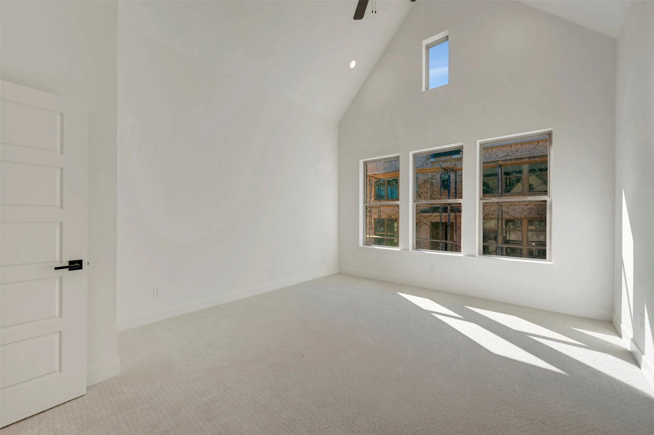 Vaulted ceiling with an accent window, three vertical windows, white paneled door with matte black hardware, and neutral carpet flooring