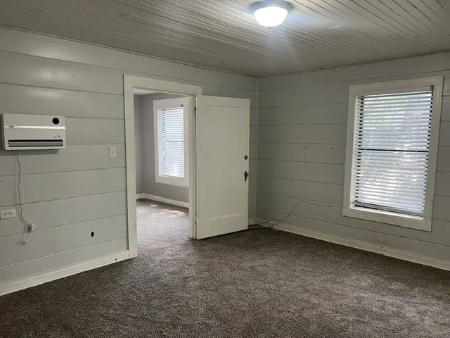 Bright and spacious room featuring neutral shiplap wall paneling, carpet flooring, and abundant natural light from a large window with plantation shutters