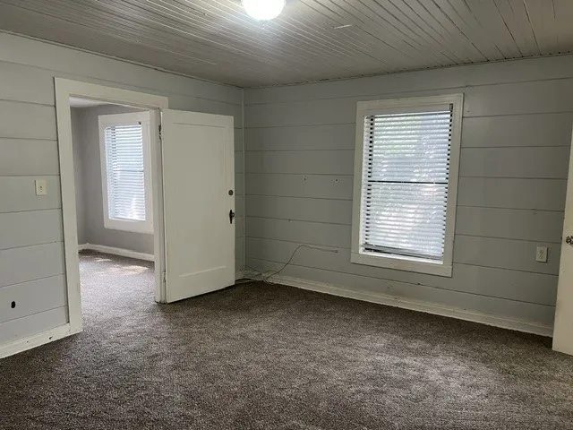 Spacious interior room featuring neutral shiplap walls, carpeted flooring, large double-hung window with blinds, and multiple doorway openings