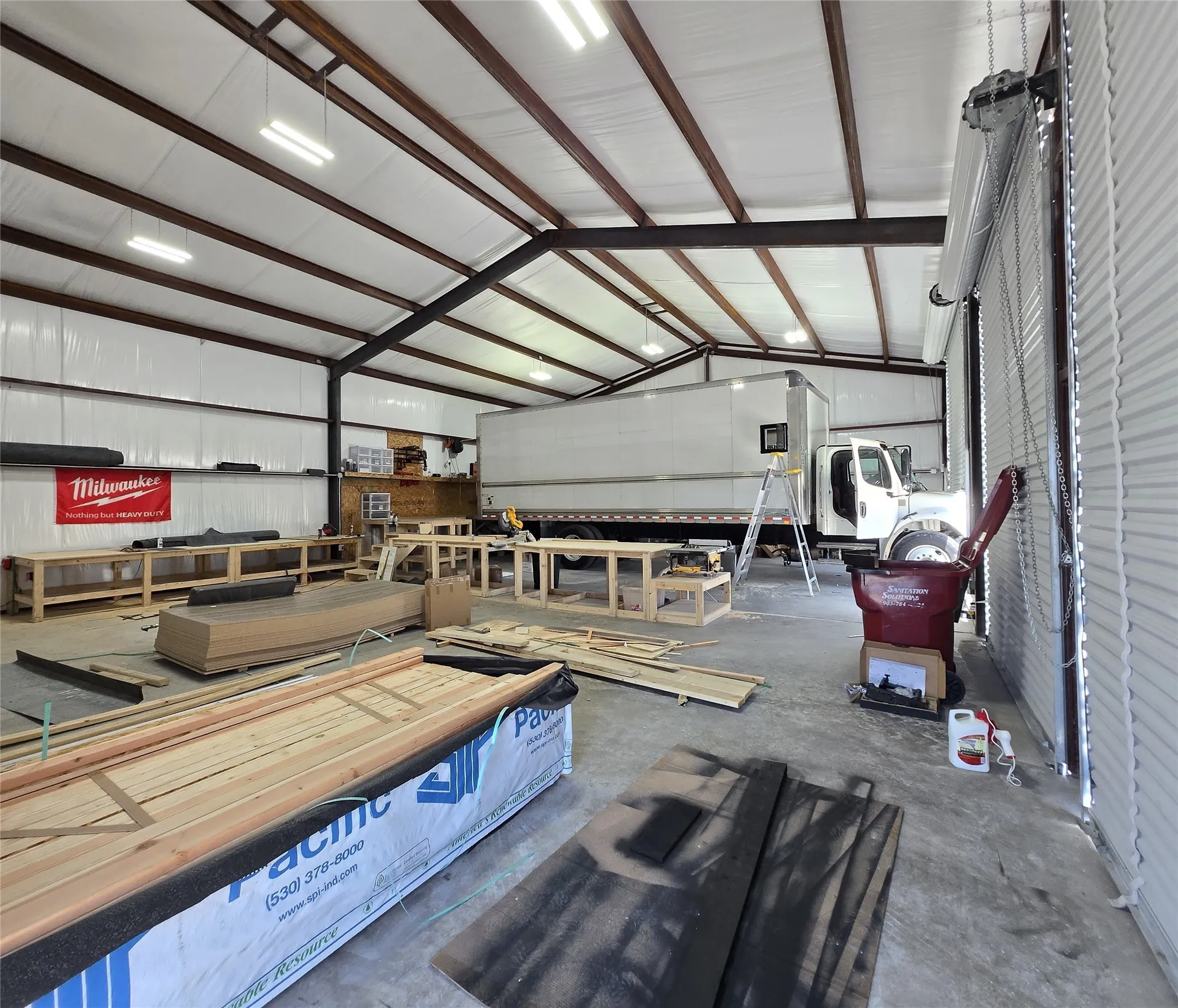 Spacious industrial interior featuring a high ceiling, visible structural beams, multiple overhead fluorescent light fixtures, a concrete floor, and a large roll-up door