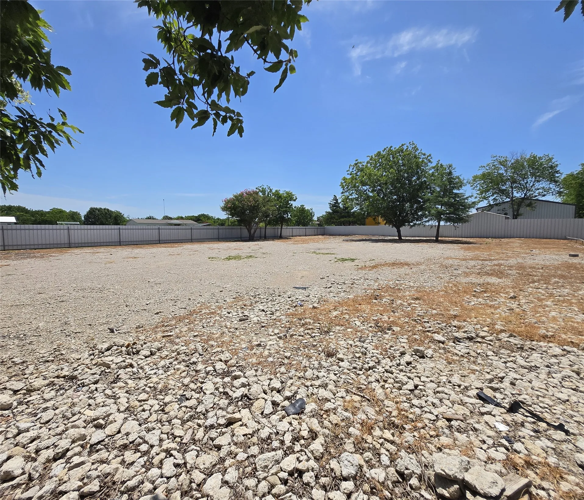 Expansive land parcel featuring a gravel surface, established trees, and a perimeter fence