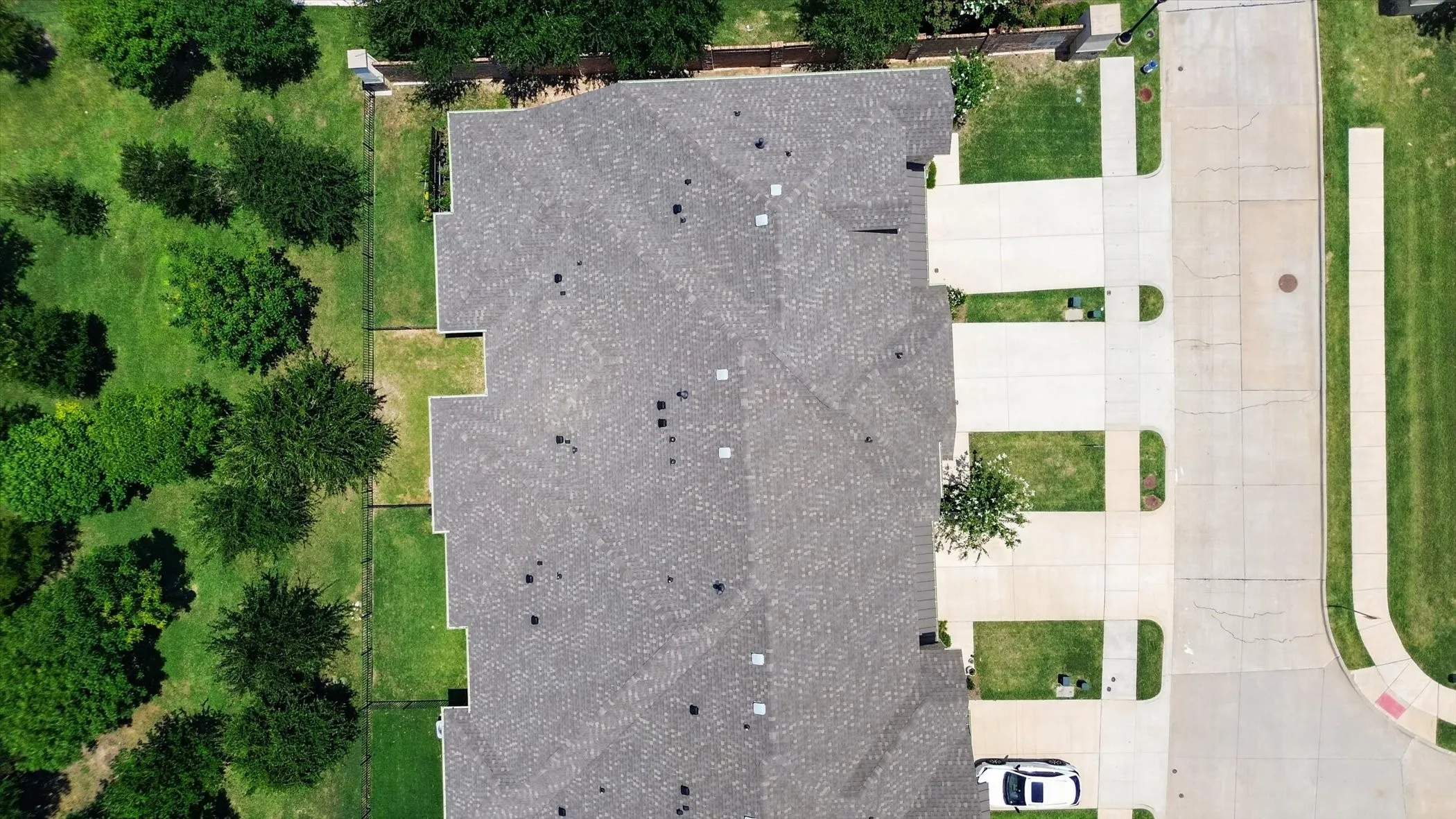 Roofline of a property with a gray shingle roof, bordered by a green landscaped area featuring mature trees and a manicured lawn