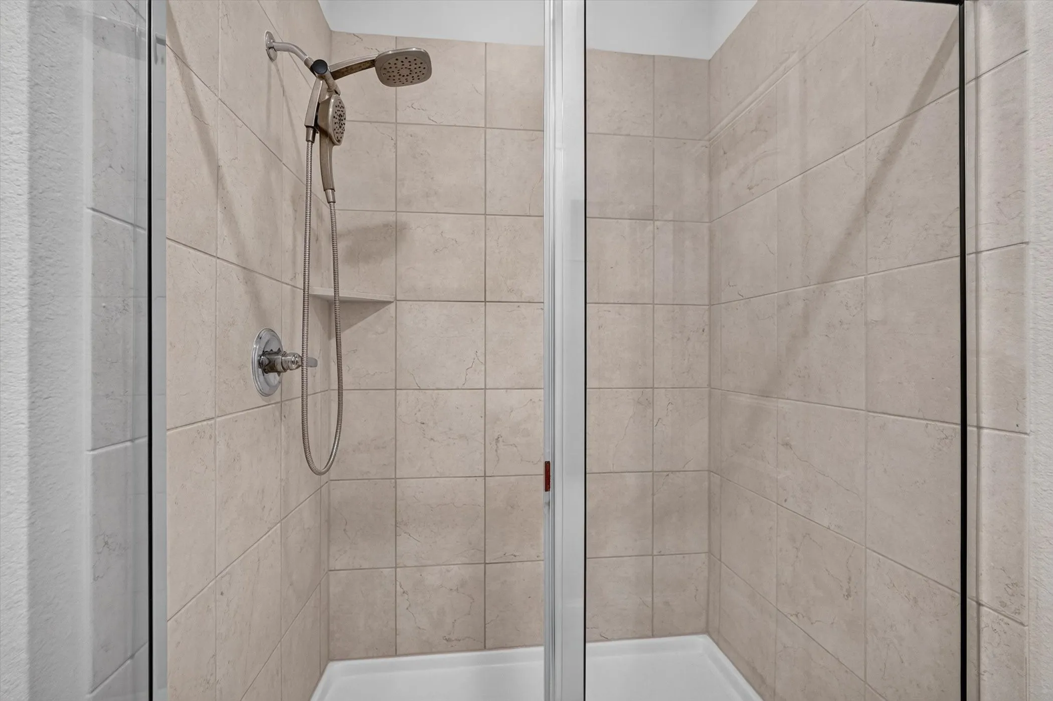 Shower enclosure with light-toned rectangular wall tiling, a white shower pan, and a brushed nickel multi-function showerhead with a handheld sprayer