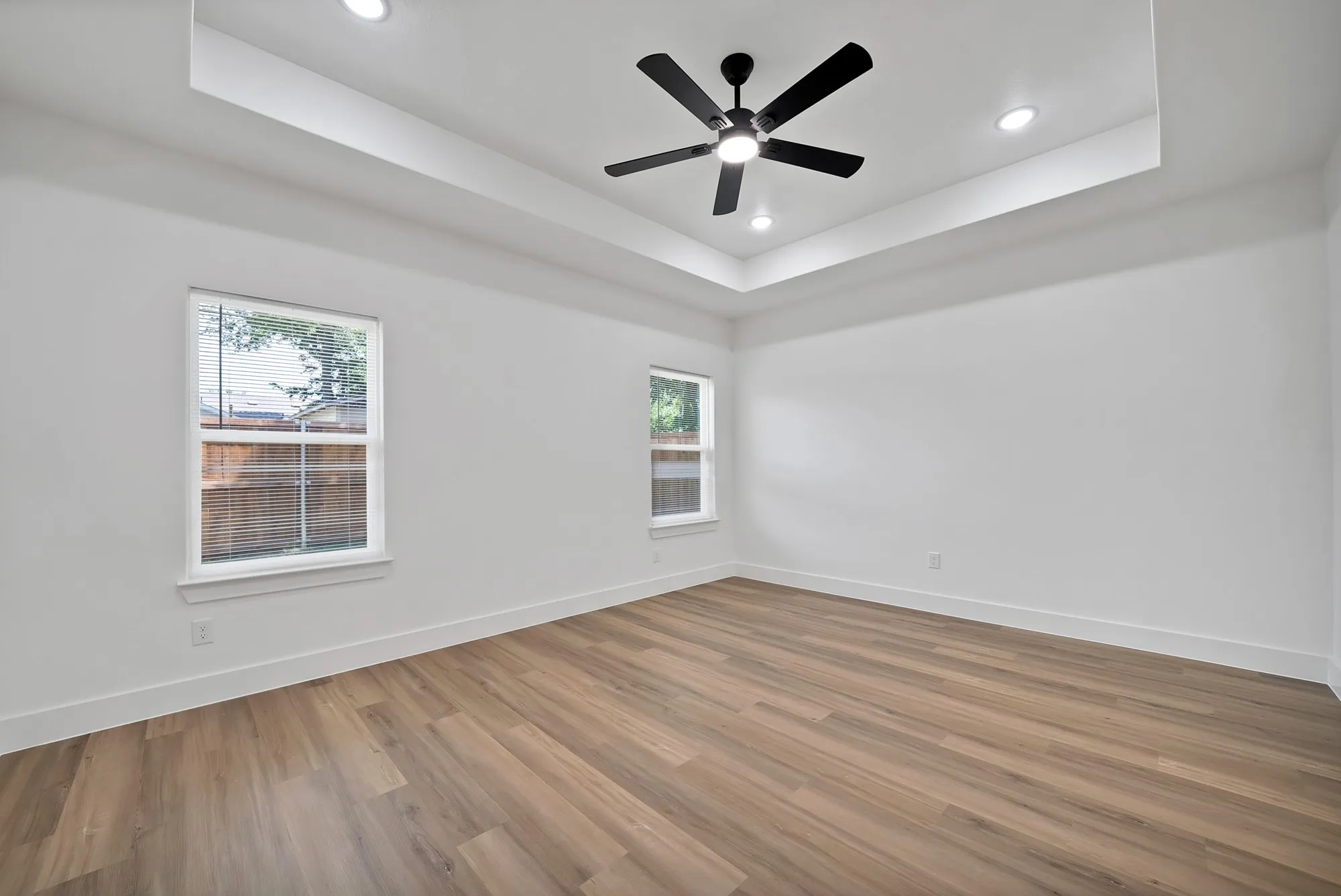 Spacious room featuring wood-finish flooring, two windows with white trim, a tray ceiling, recessed lighting, and a black ceiling fan
