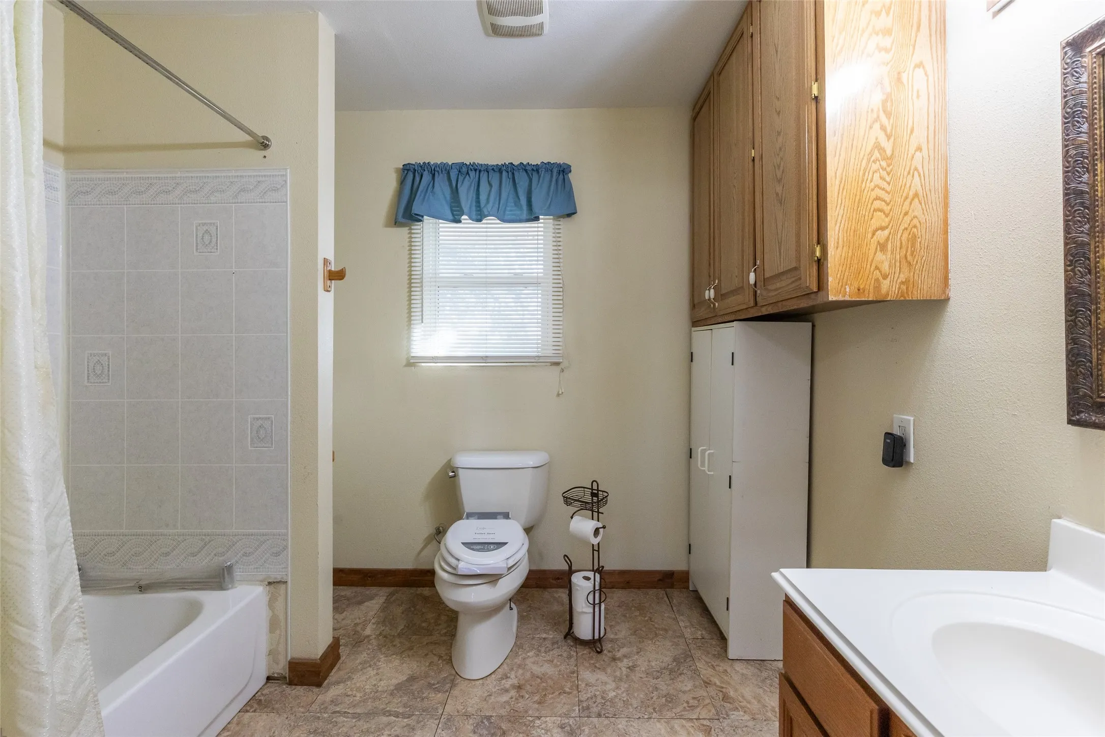 Bathroom featuring a white tub with tiled surround, a toilet, an opaque window, a vanity with an integrated sink, and wood-finish cabinetry