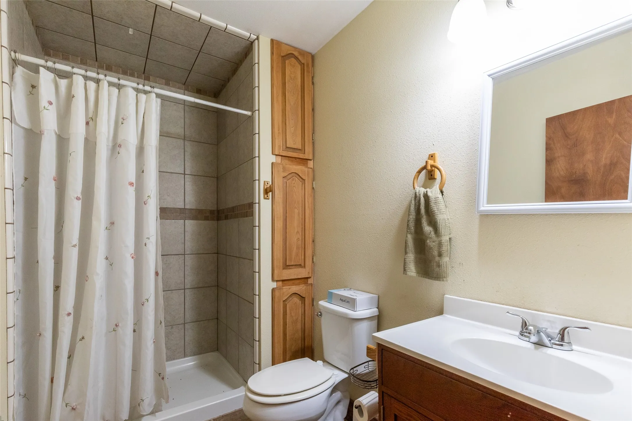 Full bathroom featuring a tiled shower with a decorative accent band, a single vanity with a white countertop and integrated sink, a large wall-mounted mirror, and wood-finish cabinetry