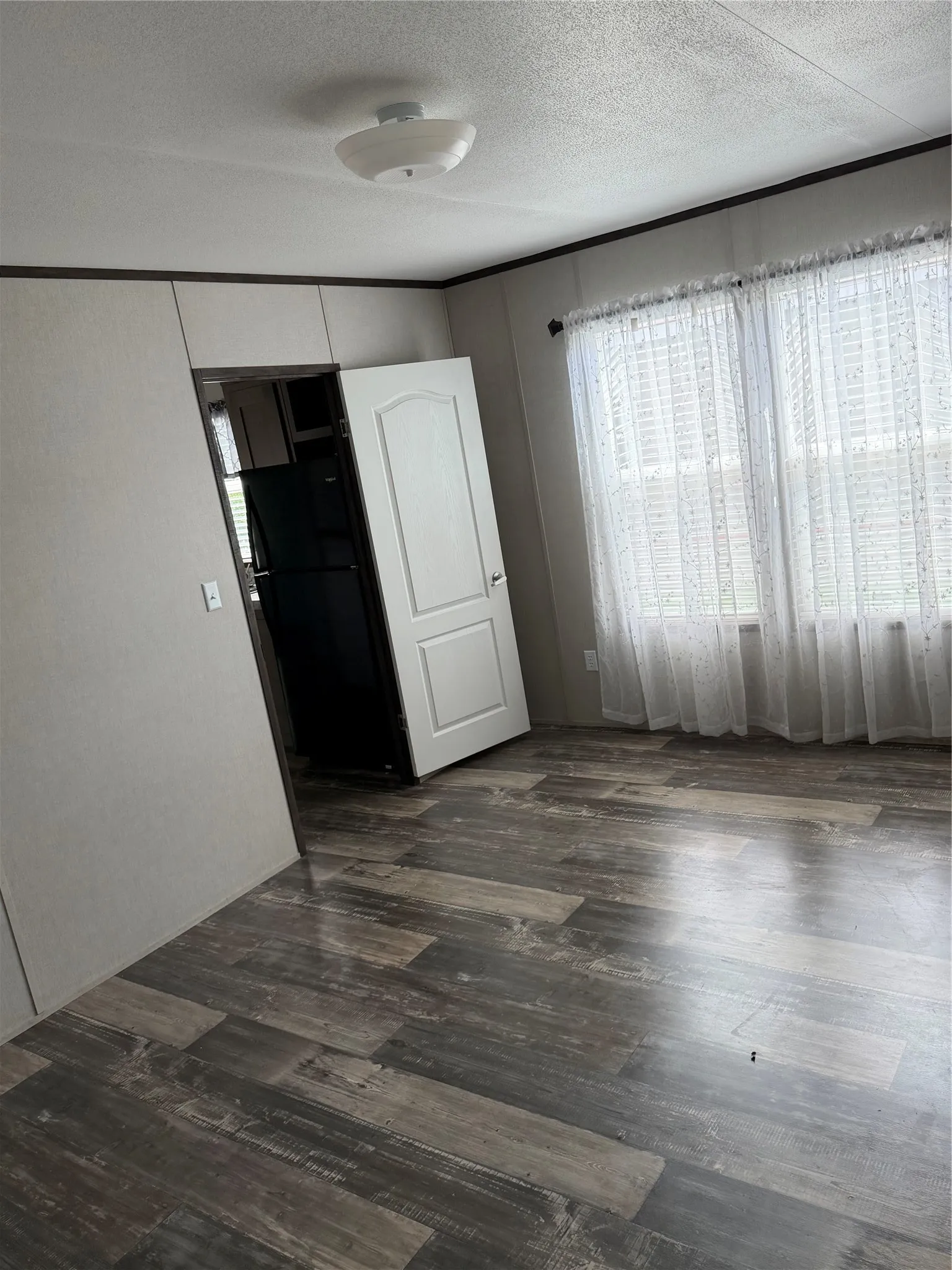 Spacious room featuring wood-finish flooring, white walls with dark trim, and a bright window with sheer curtains