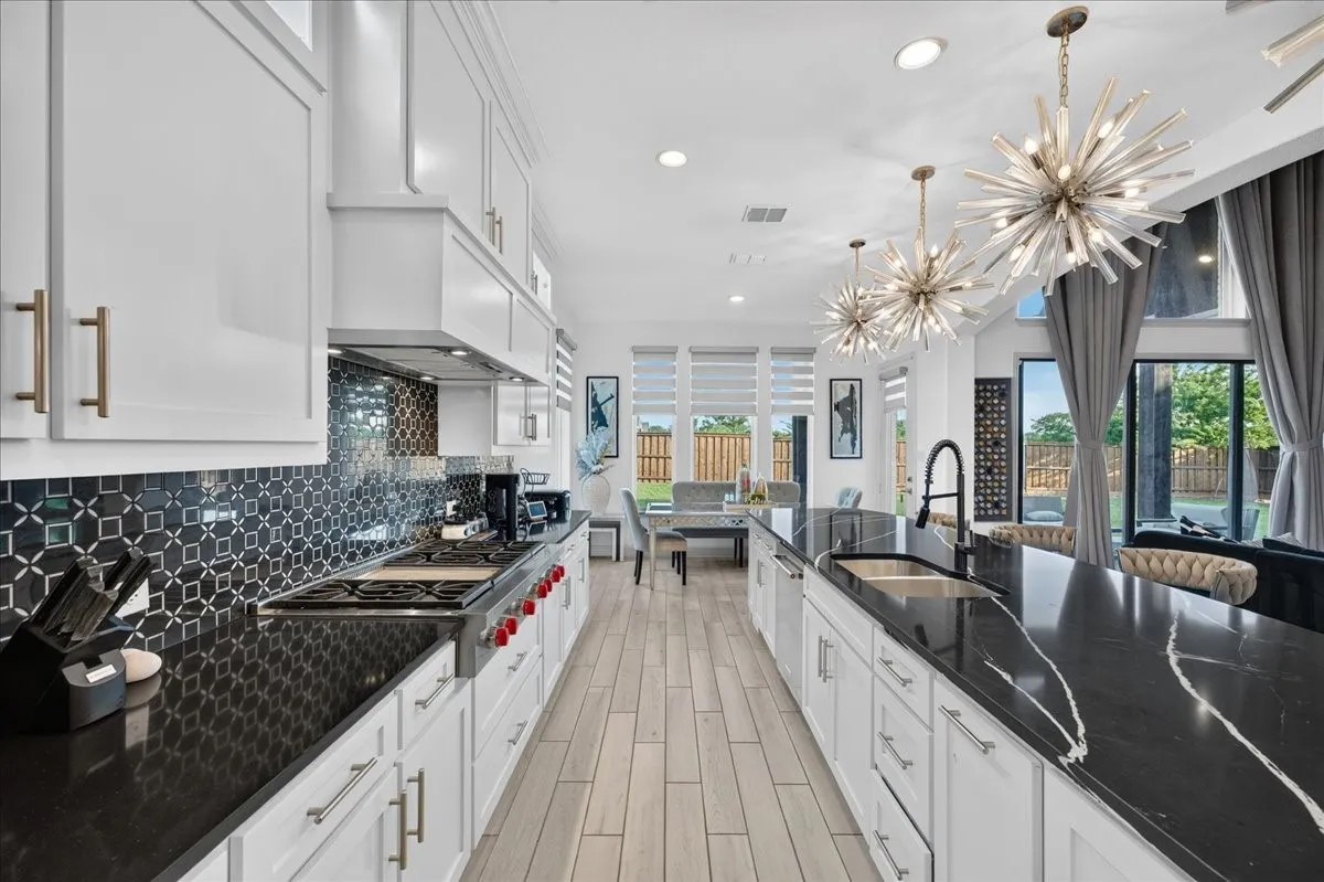 Spacious kitchen featuring an elongated island with integrated sink, black countertops with white veining, white cabinetry, a patterned black tile backsplash, and light wood-finish flooring