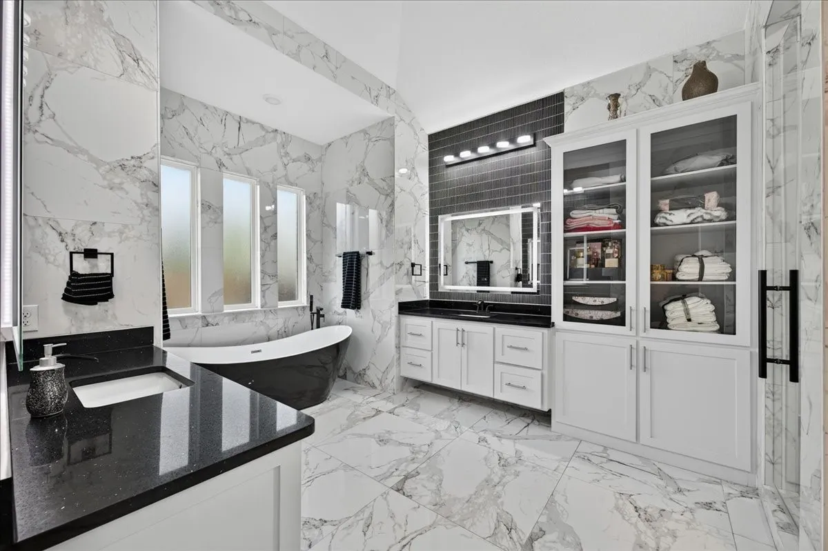 Spacious bathroom featuring marble-look tile flooring and walls, a freestanding black soaking tub, a dual vanity with dark countertops and white cabinetry, built-in storage cabinets with glass doors, and contemporary lighting fixtures