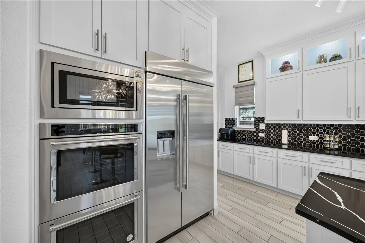 Gourmet kitchen featuring stainless steel appliances, a built-in oven and microwave, light wood-finish flooring, white cabinetry, and a black patterned tile backsplash