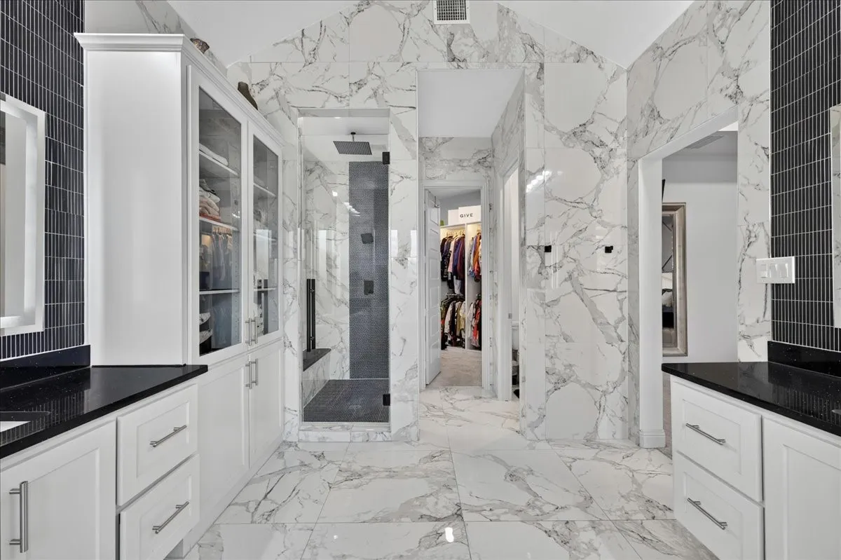 Spacious bathroom featuring white cabinetry with dark countertops, a walk-in shower with a glass enclosure, white marble-look tile flooring, and white marble-look tile walls