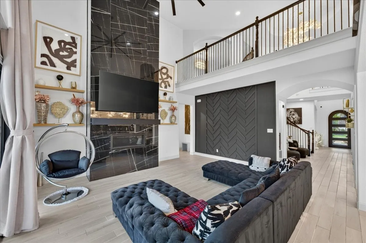 Two-story living area featuring a dramatic black stone-finish fireplace, light wood-finish flooring, a contemporary accent wall with chevron paneling, and an upper-level balcony with black iron railings