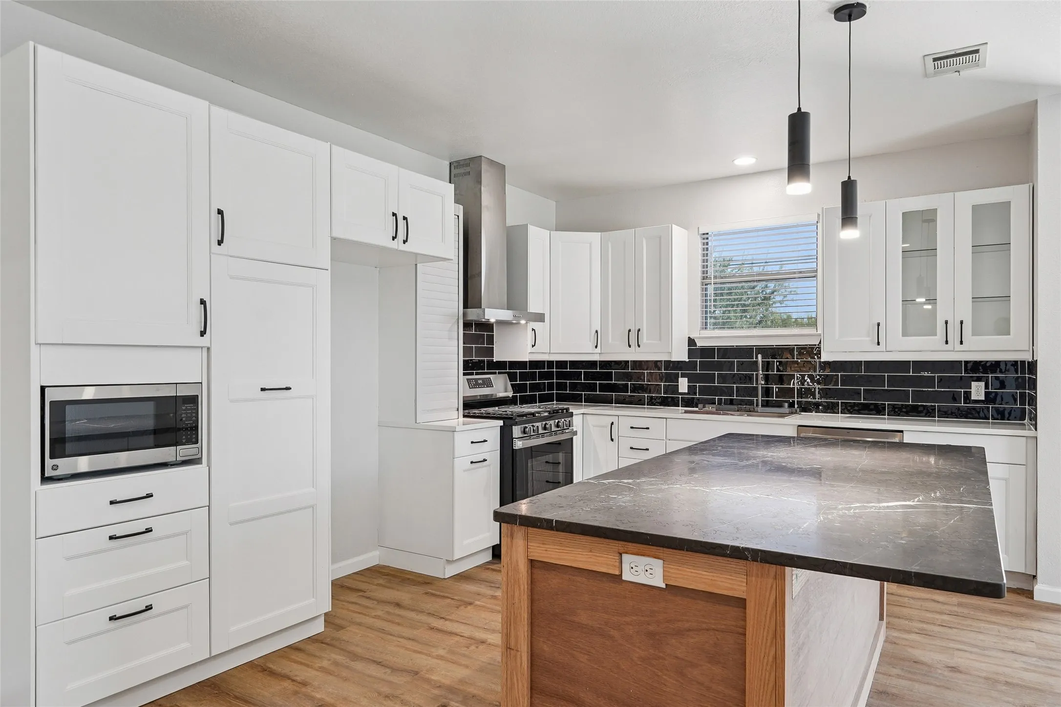 Contemporary kitchen featuring white cabinetry, a dark stone island countertop, a black subway tile backsplash, stainless steel appliances, and wood-finish flooring