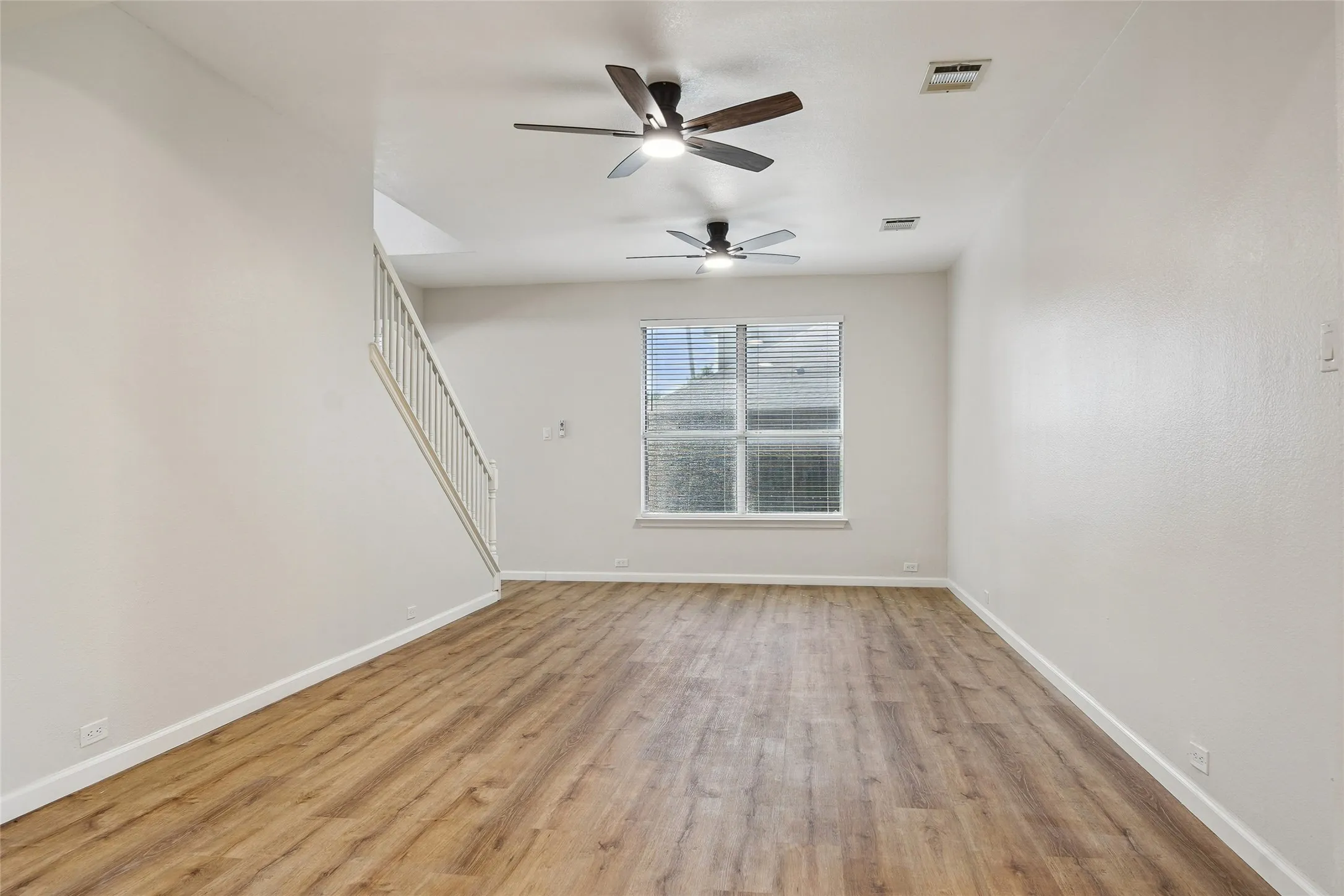 Wood-finish flooring, white walls, two ceiling fans with lighting, window with blinds, and staircase with white railing